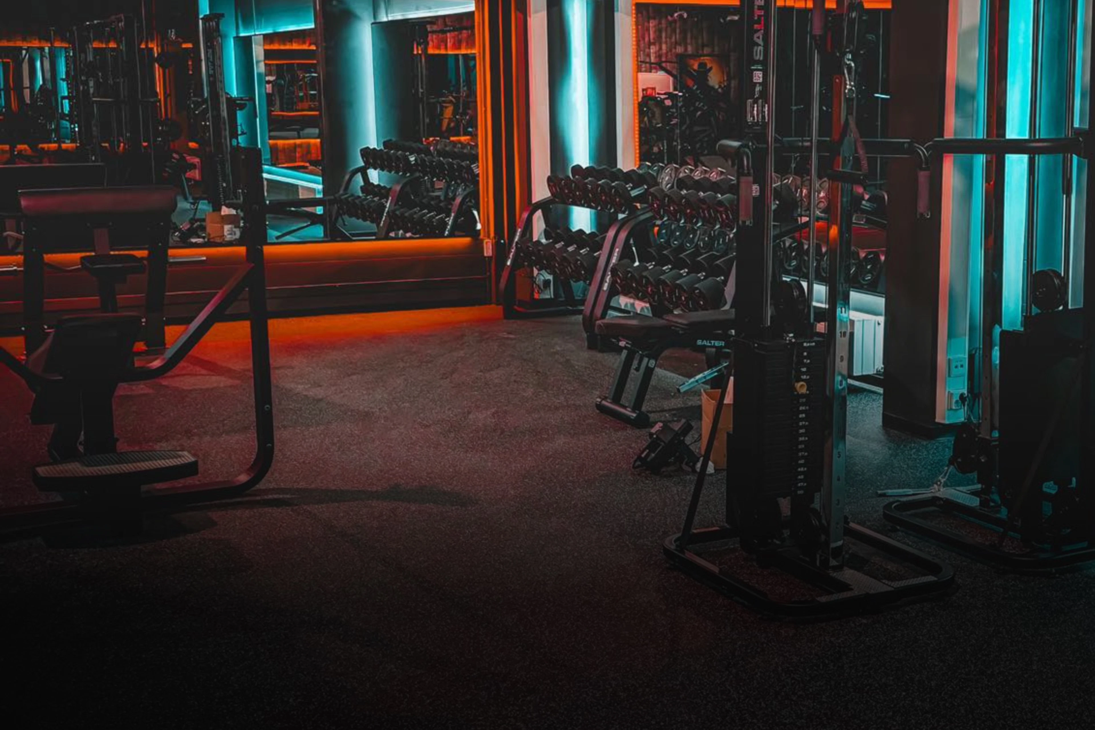Modernt gym med färgglad blåorange belysning. Synliga hantelställ, styrkemaskiner och speglande väggar. Gymmet har jämn golvyta.