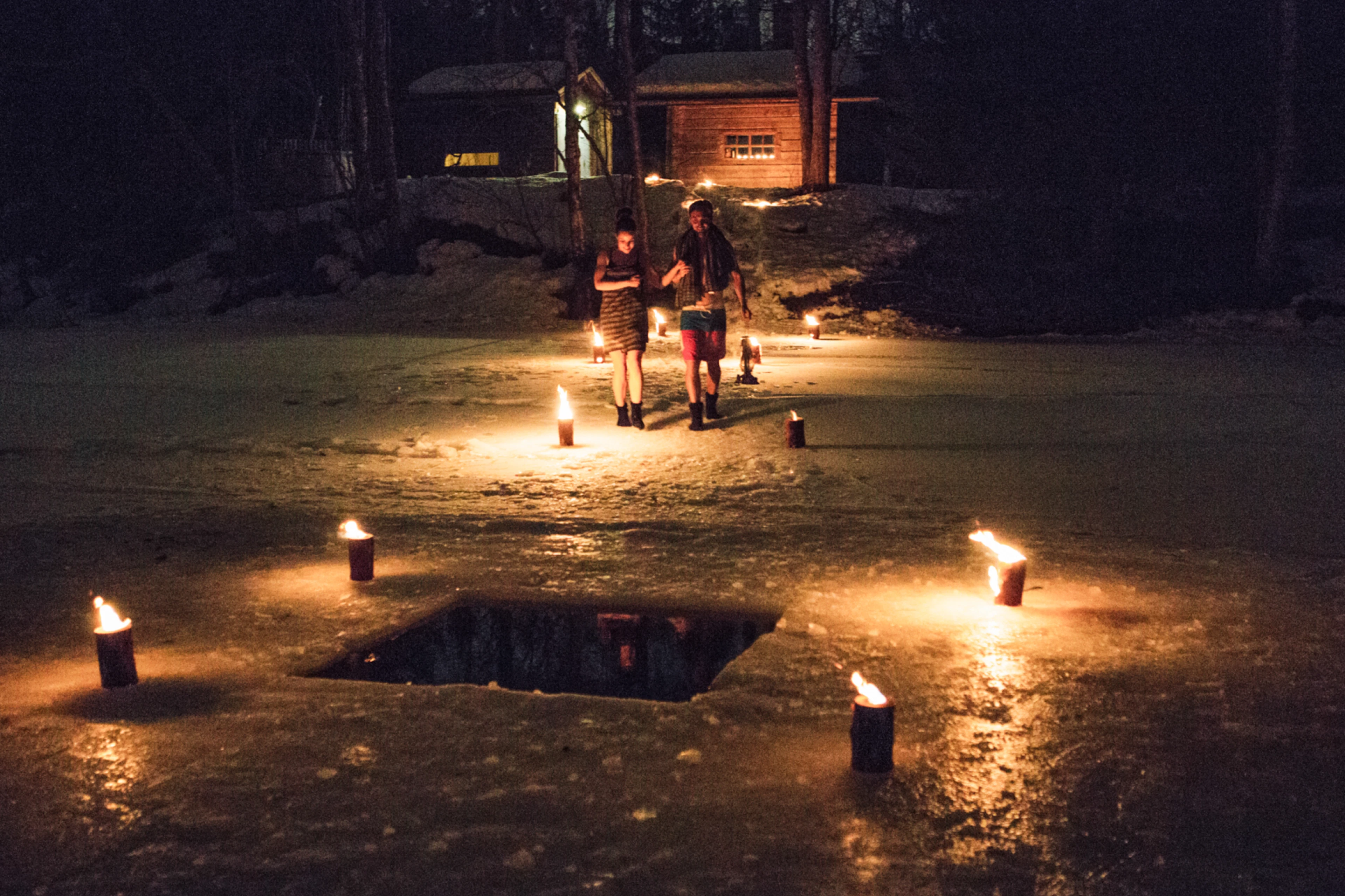 Öinen avanto jäässä, jota valaisevat tulet. Kaksi henkilöä seisoo jään päällä ja taustalla näkyy puinen saunamökki metsässä.