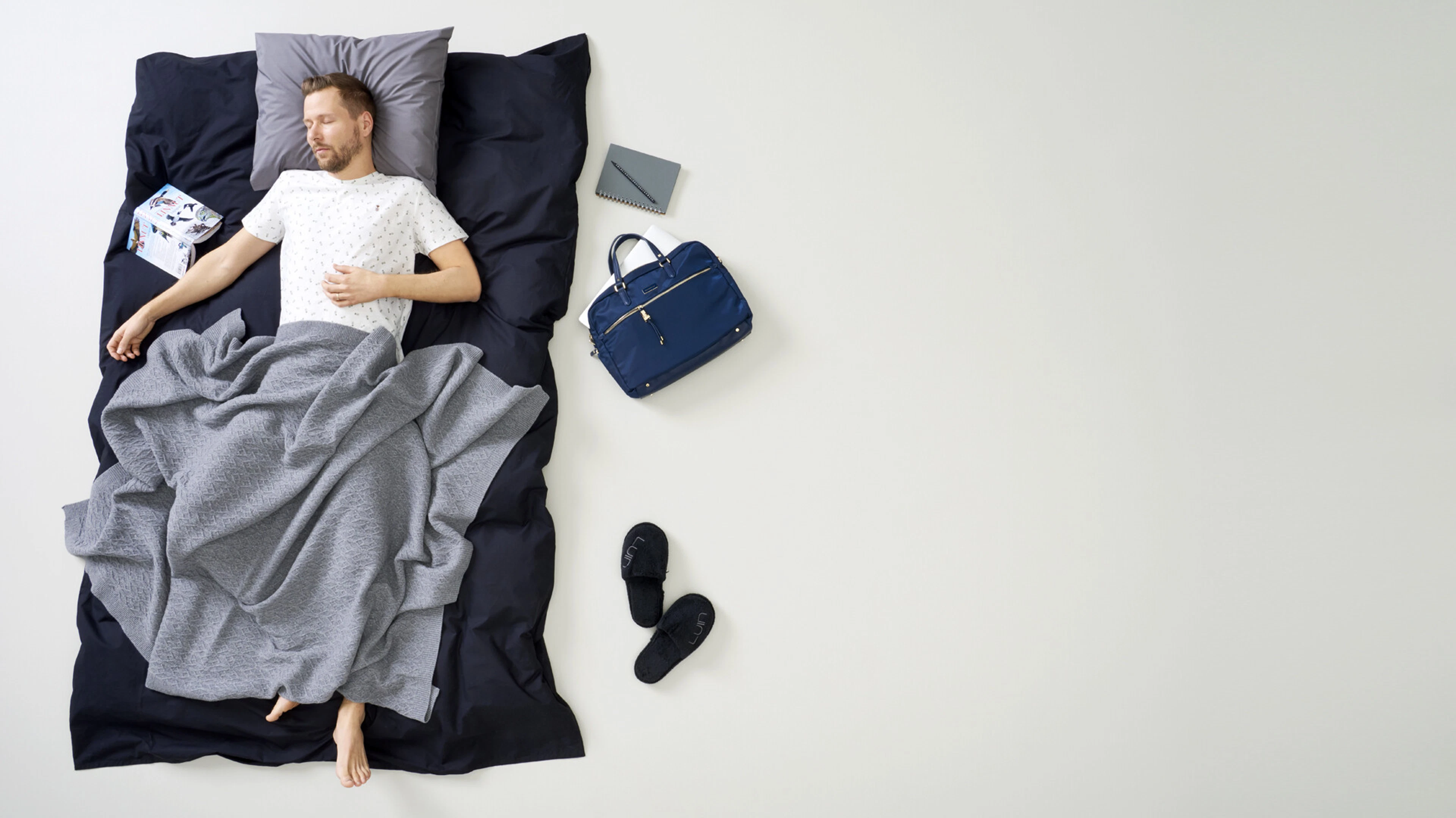 Man sover i säng med väska, sandaler och anteckningsbok på golvet bredvid.