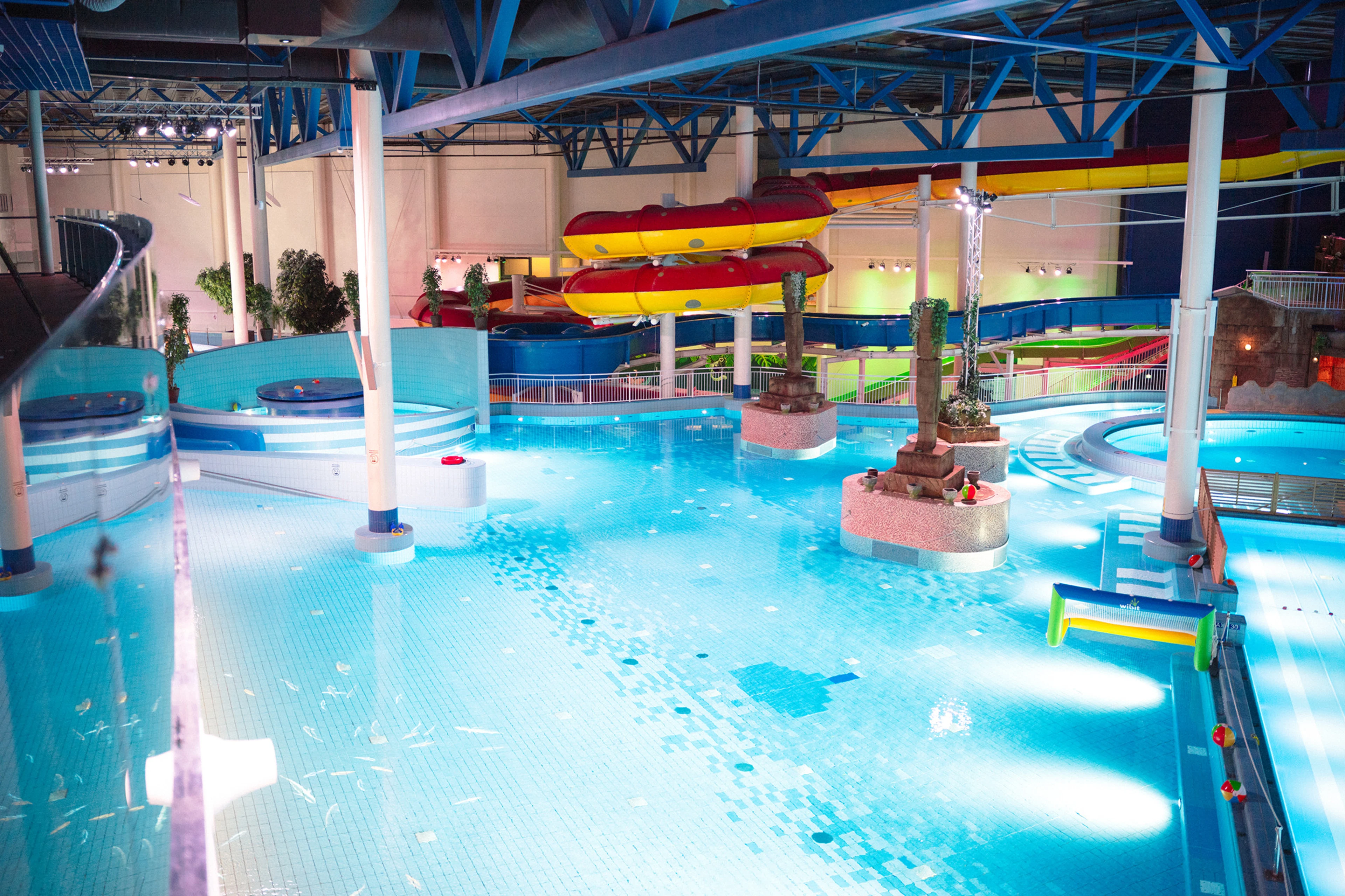 Indoor water park with blue pools, red and yellow water slides, and decorative stone pillars. Lighting enhances the blue water tones.