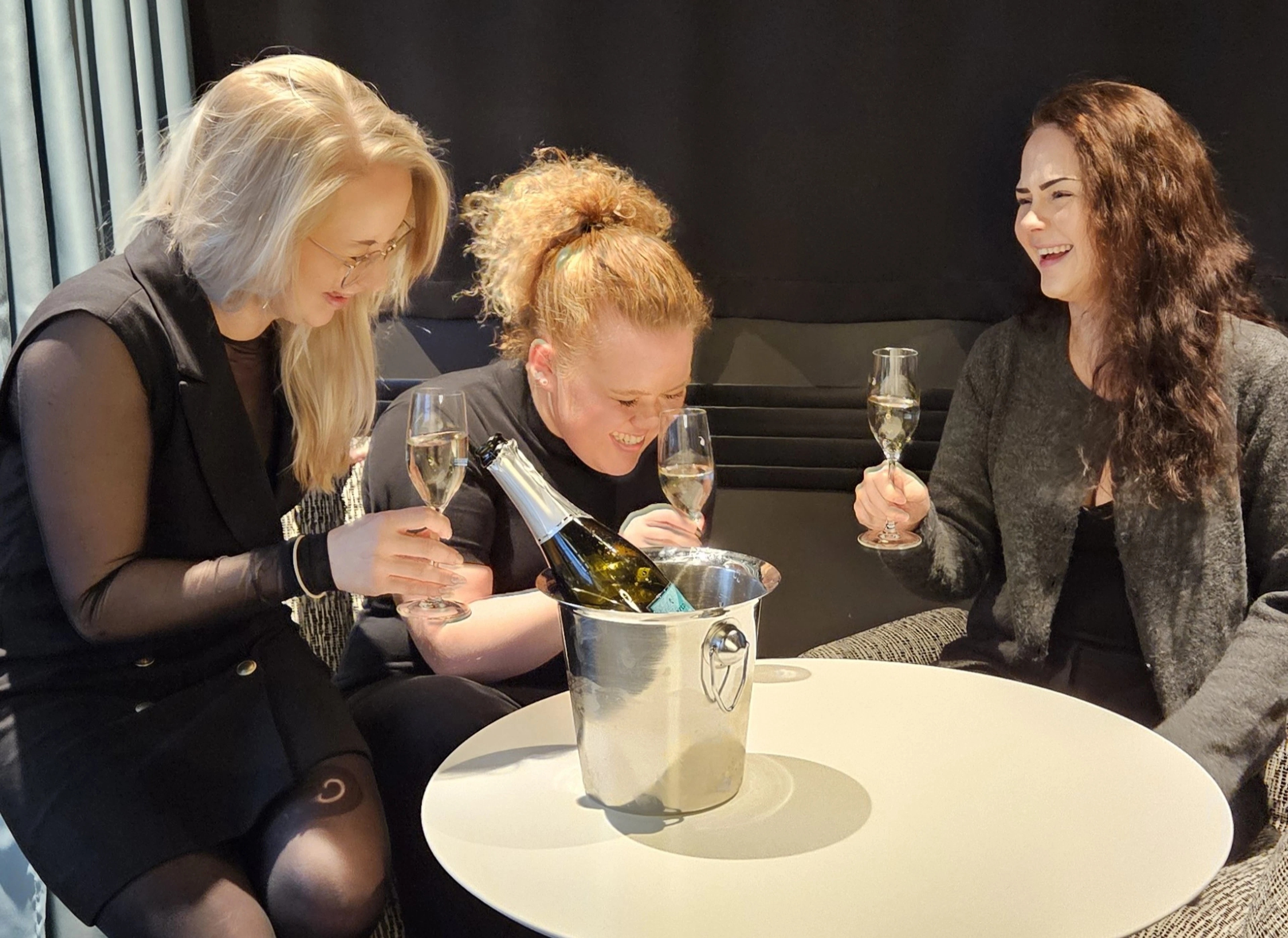Three friends laughing in a hotel room with champagne glasses in hand.