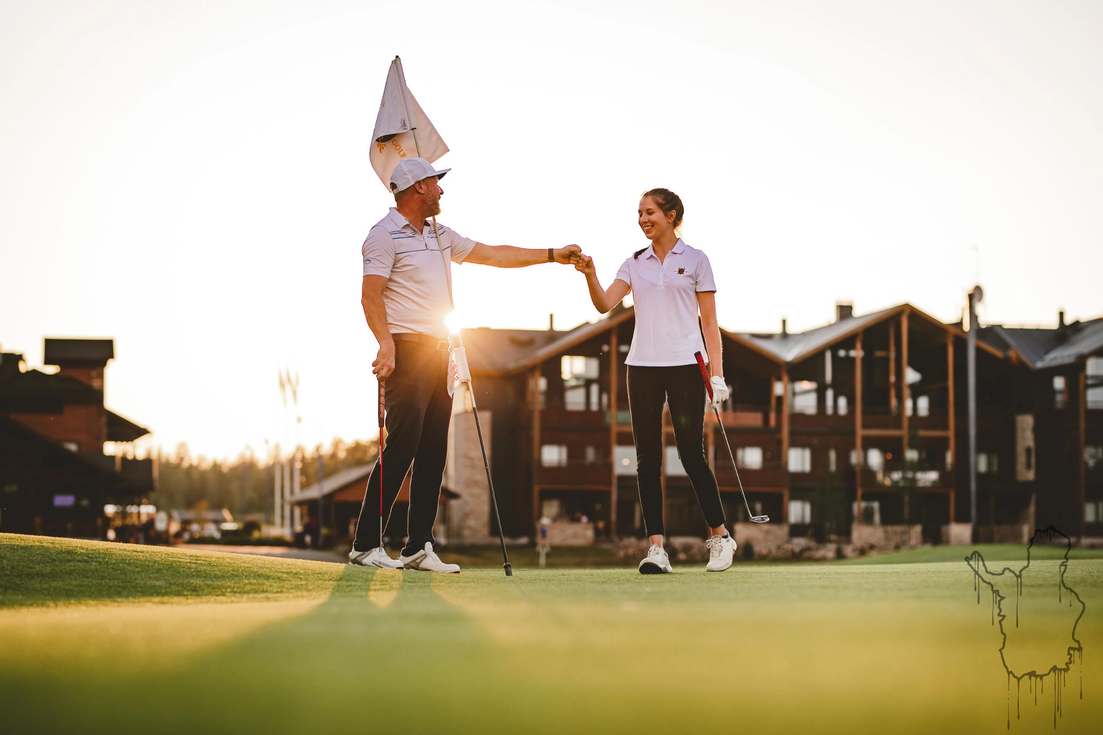 Two golfers shaking hands on putting green at sunset, low-rise buildings in background.