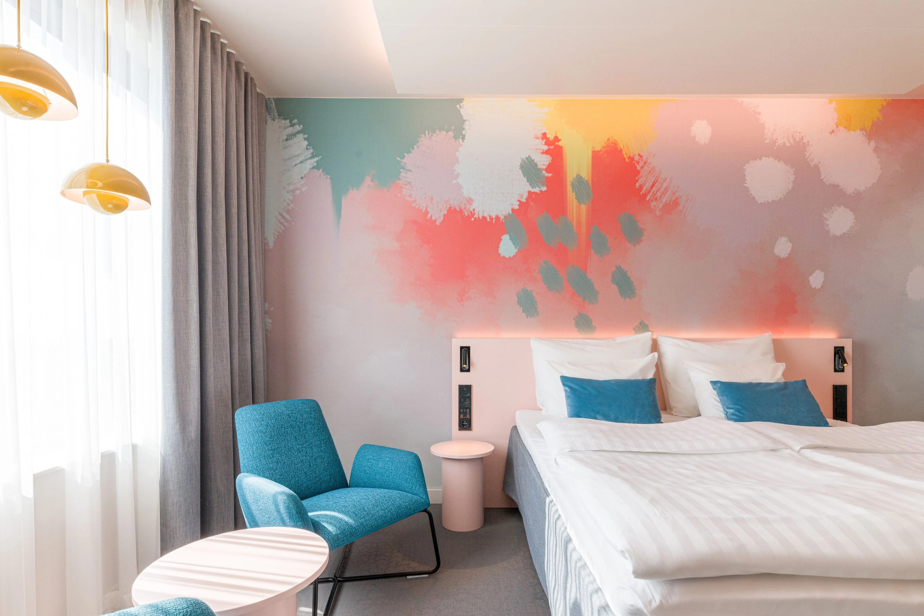 A room with a bed for two people and next to it a table with two chairs. Behind the bed there is bright colourful abstract mural wall.