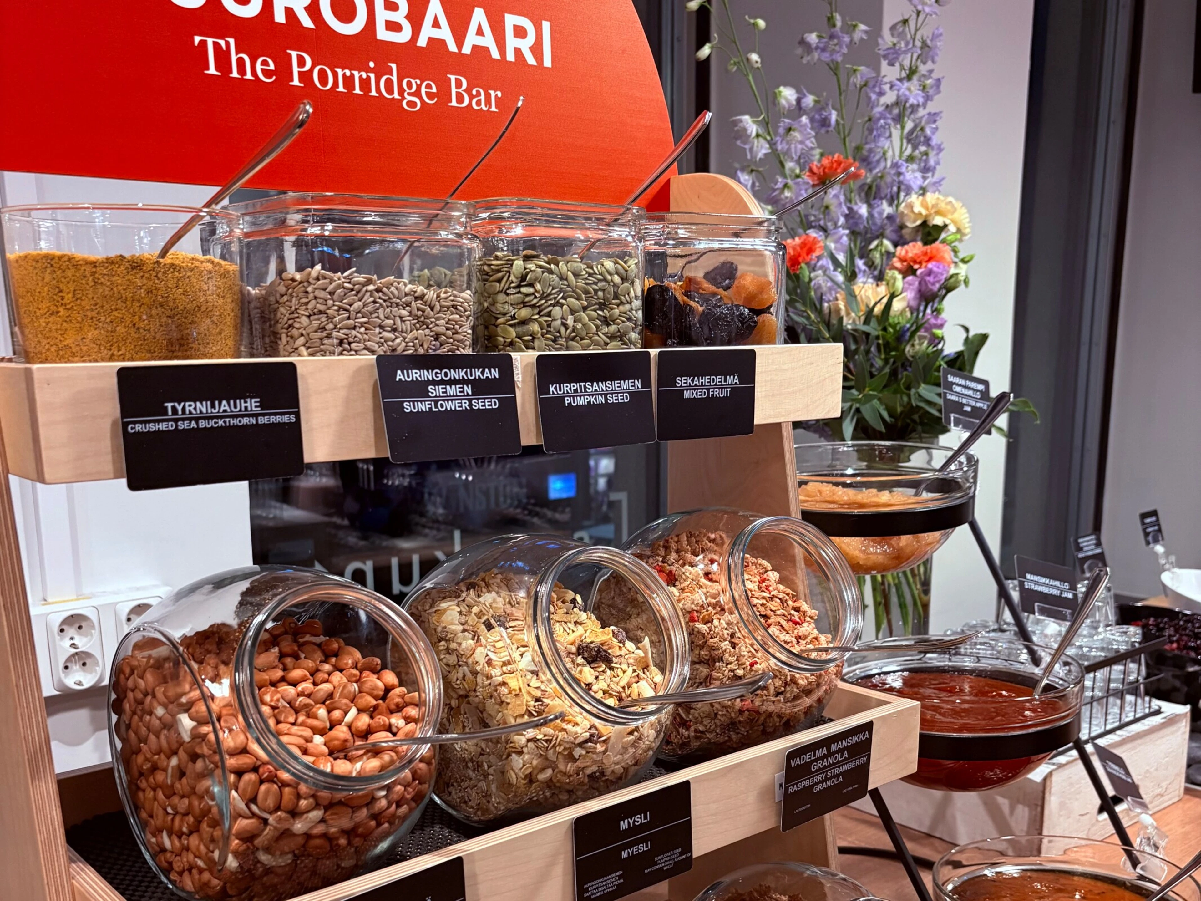 Grötbar i trä med glasburkar fyllda med frön, torkad frukt, nötter, müsli och granola. Intill finns sylt i skålar och en blombukett.