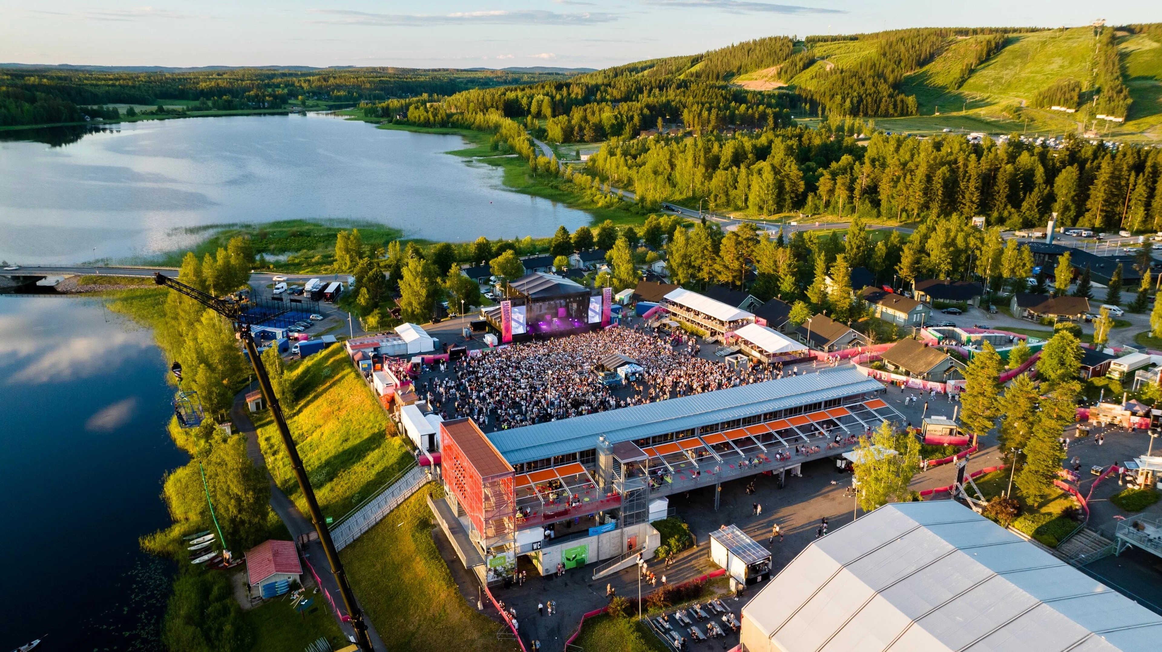 Festivaalialue järven rannalla, jossa avoin päälavat, laajat asfalttialueet, väliaikaiset telttarakenteet ja metsäisiä rinteistä taustalla.