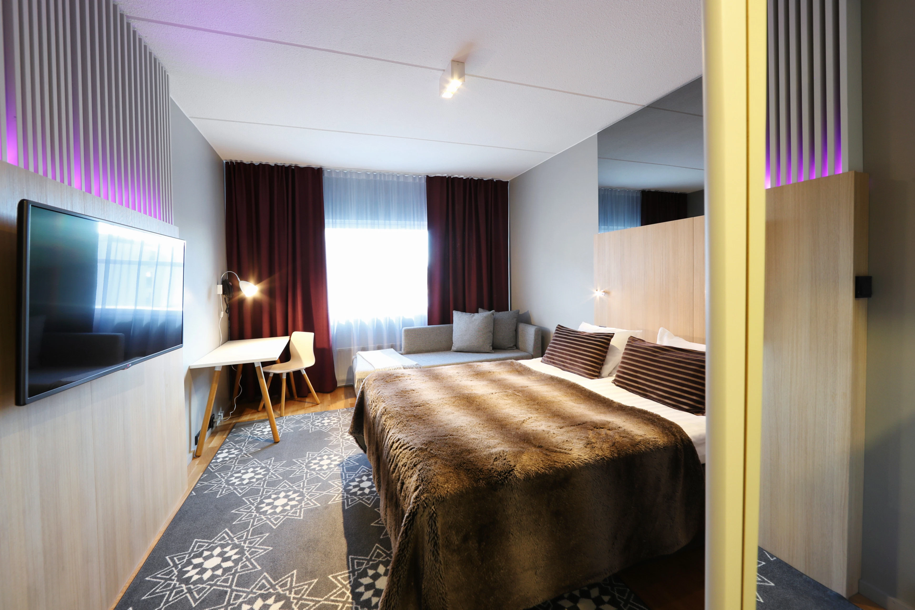 Moderni hotellihuone, jossa on suuri sänky, sohva, työpöytä, televisio seinällä ja punaruskeita verhoja. Huoneessa on puulattia ja kuvioitu matto.