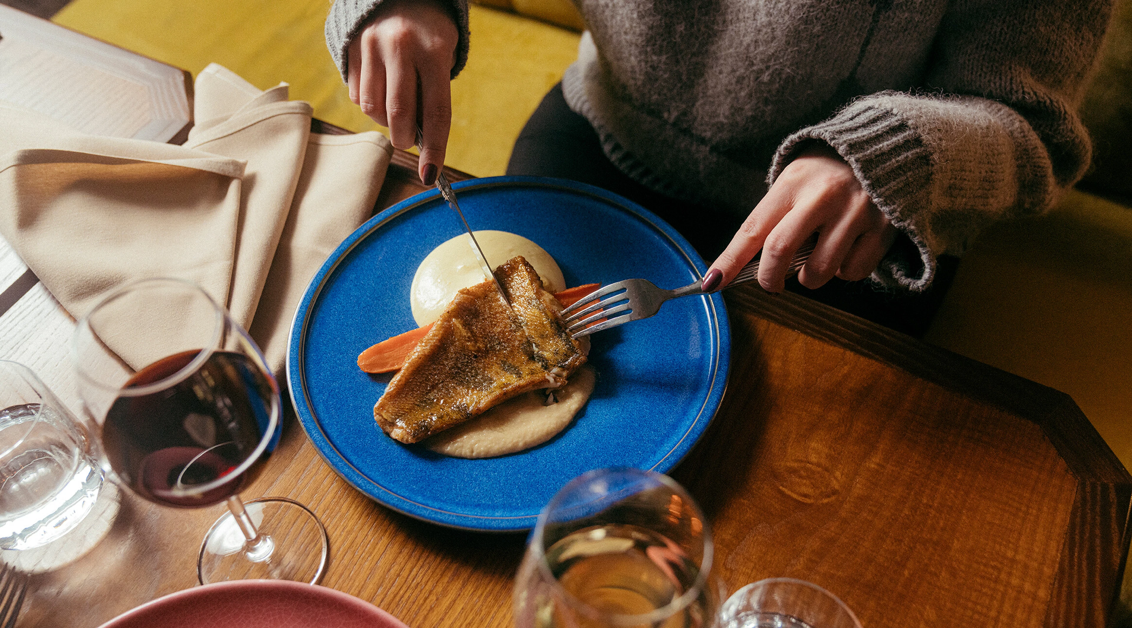 Person cutting pan-fried fish fillet with knife and fork on a blue plate at a wooden table in a restaurant.