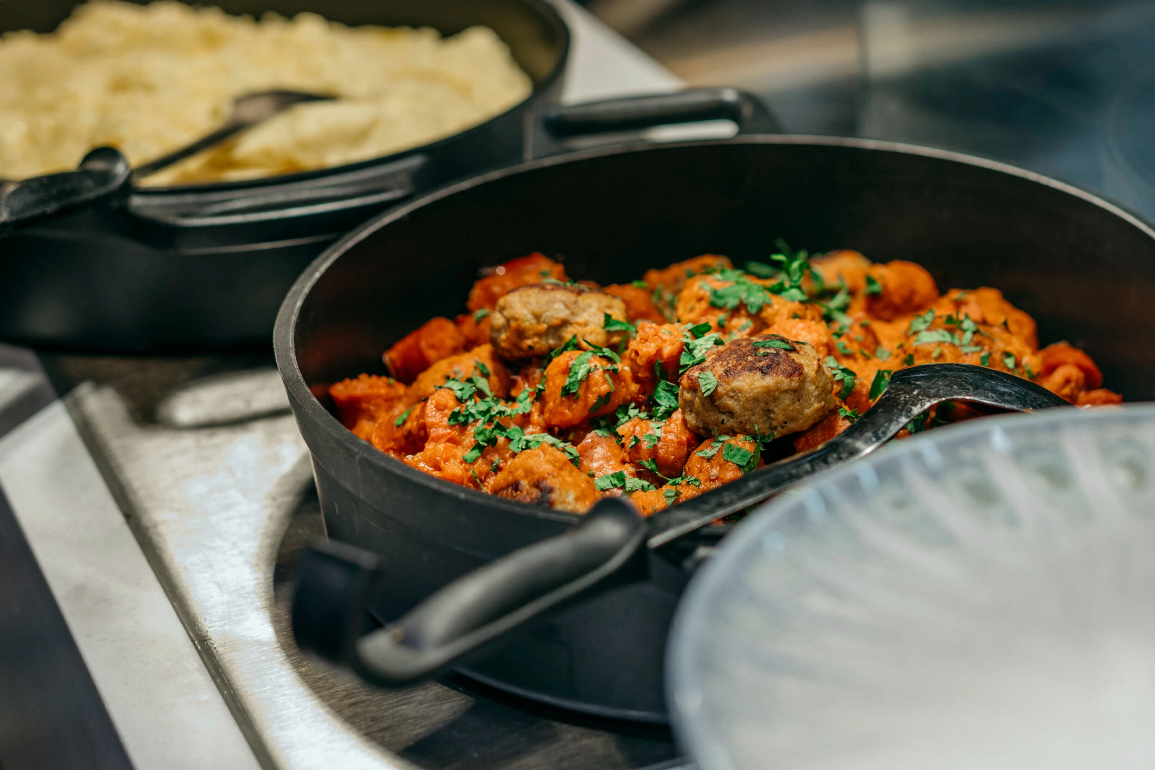 Cast iron pan with meatballs in tomato sauce garnished with fresh herbs. A porridge pot visible in the background.