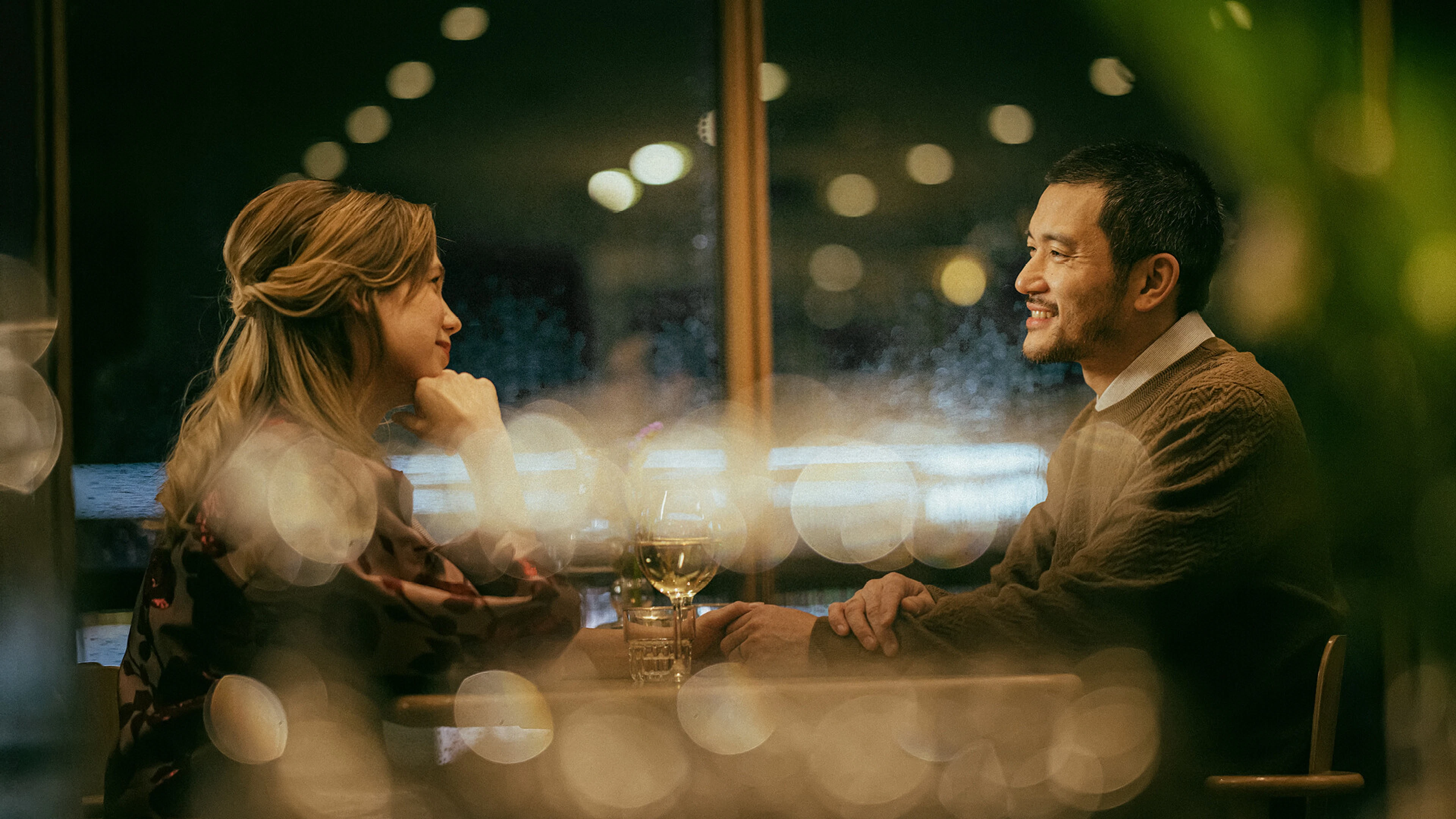 Two people conversing at a restaurant table. The space has dim lighting and large windows.