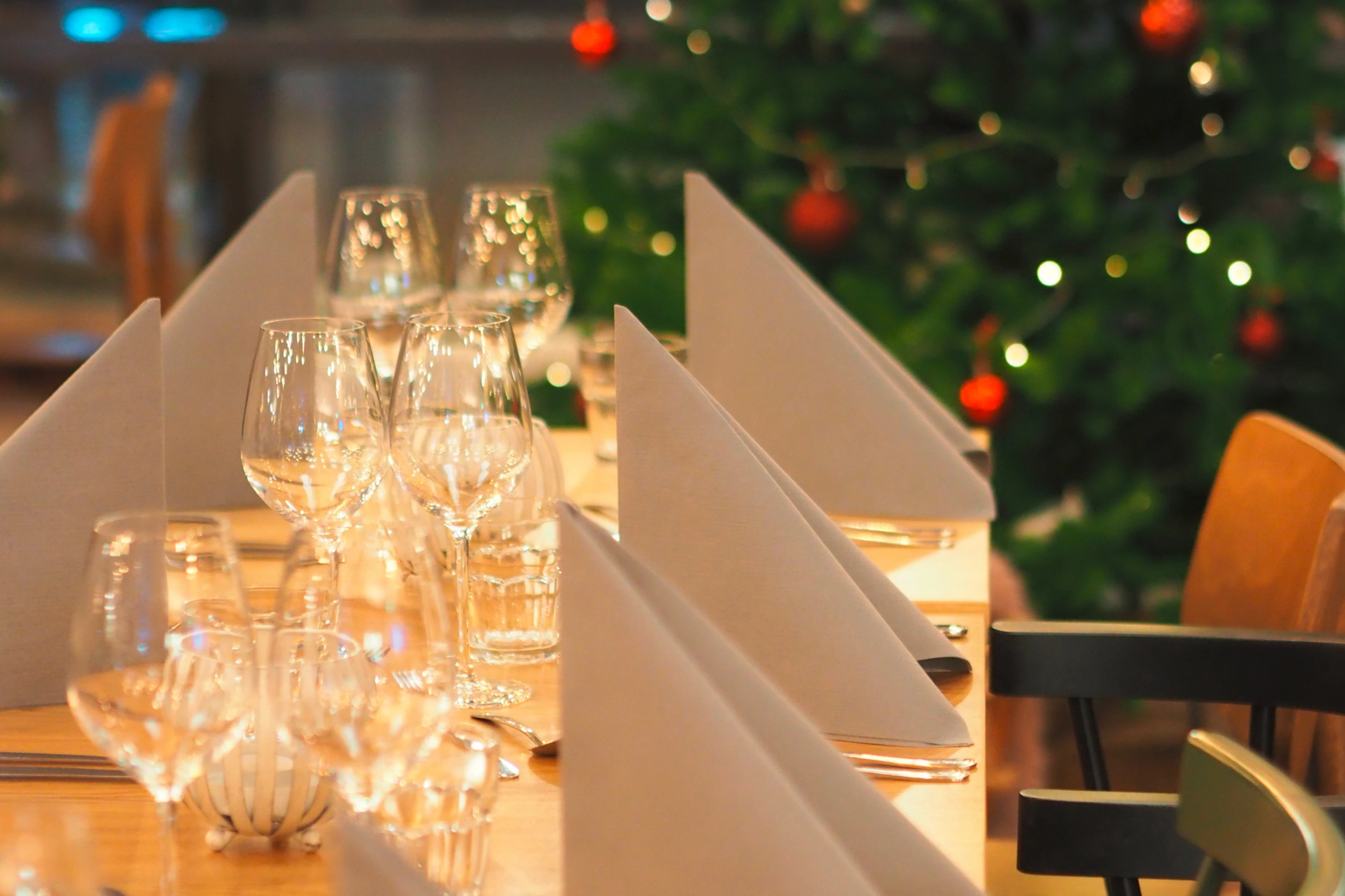 Tyylikkäästi katettu ravintolan pöytä, jossa on viinilaseja ja valkoisia lautasliinoja. Taustalla näkyy koristeltu joulukuusi.