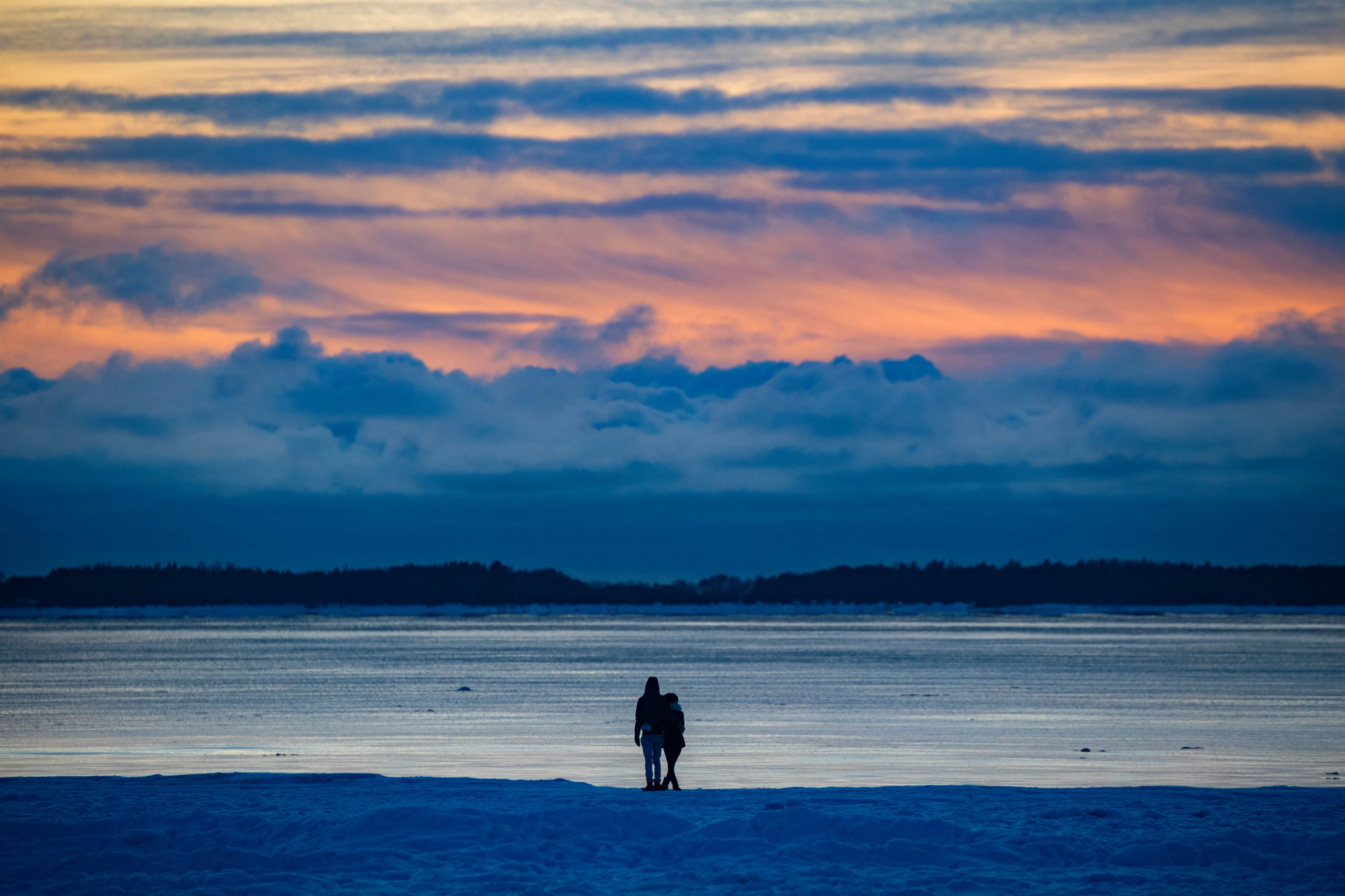 Two people standing on a snowy beach looking out at the sea, with sunset-colored clouds and a forested island in the background.
