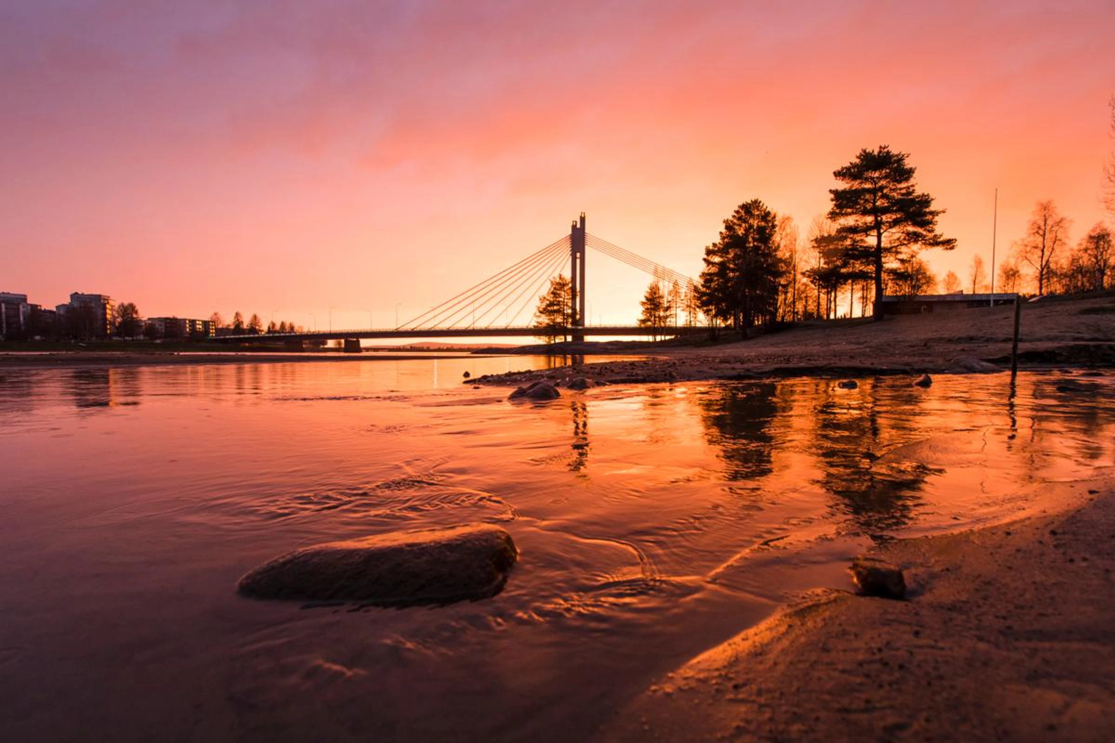 Silta joen yli auringonlaskussa, taustalla kaupunki ja etualalla vettä ja rantaviivaa. Oranssi taivas heijastuu vedenpinnasta.