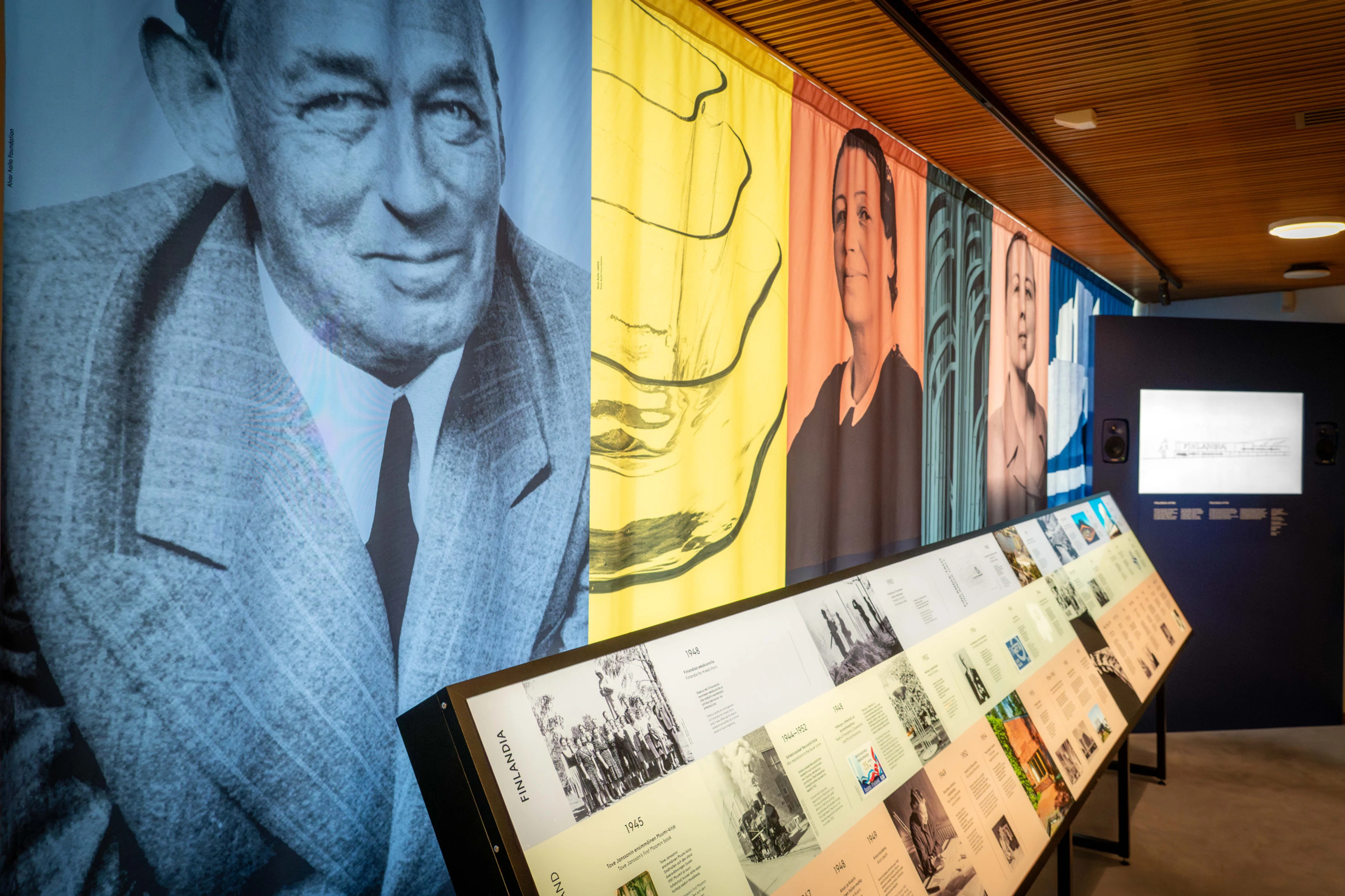 Museum exhibition space featuring large black and white portraits on the wall and colorful information panels on the lower level.