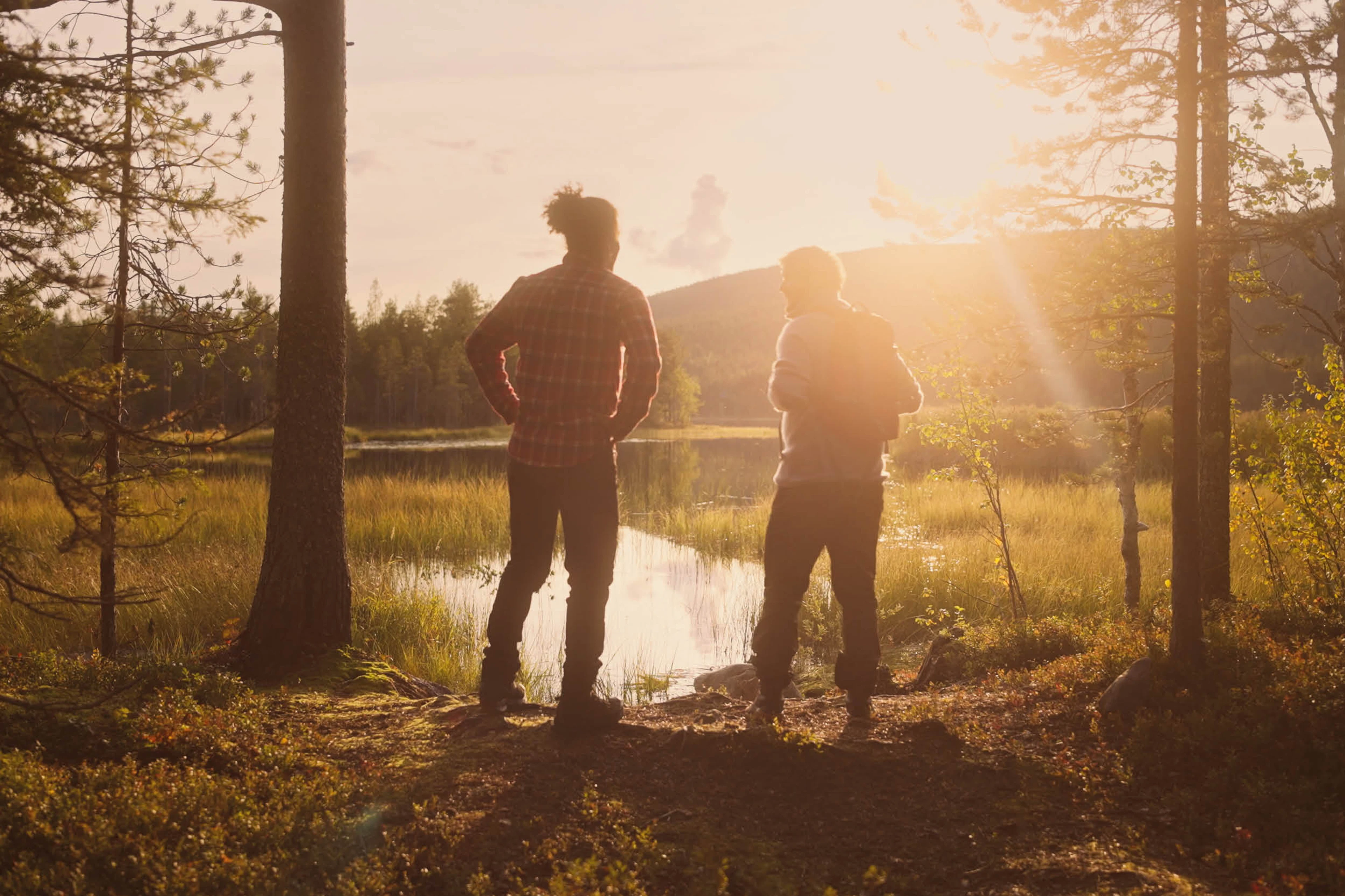 Kaksi henkilöä seisoo metsälammen rannalla auringonlaskun aikaan, kultainen valo siivilöityy puiden läpi.
