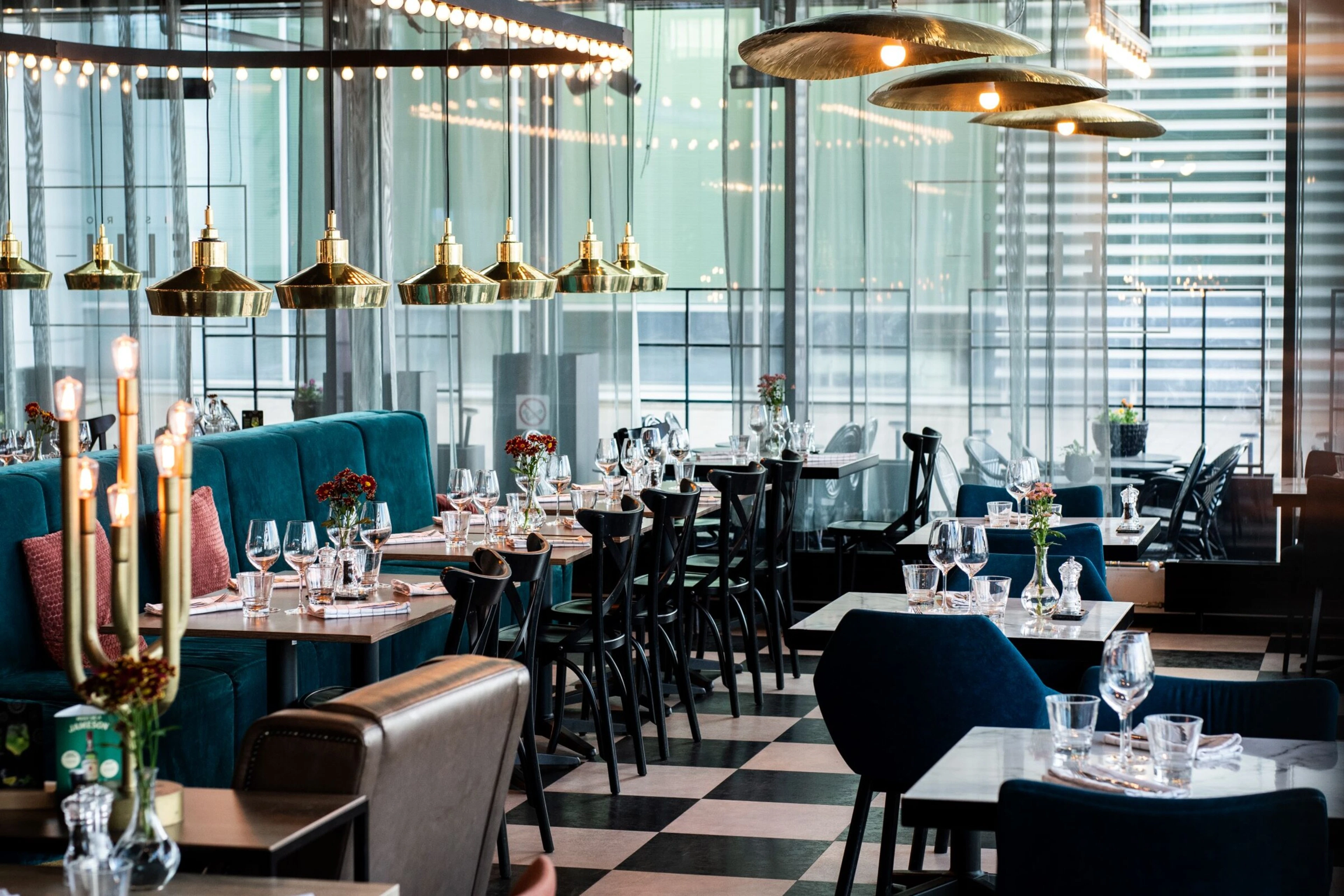 Restaurant dining room with multiple set tables, blue and black chairs, brass pendant lights, and checkered floor.