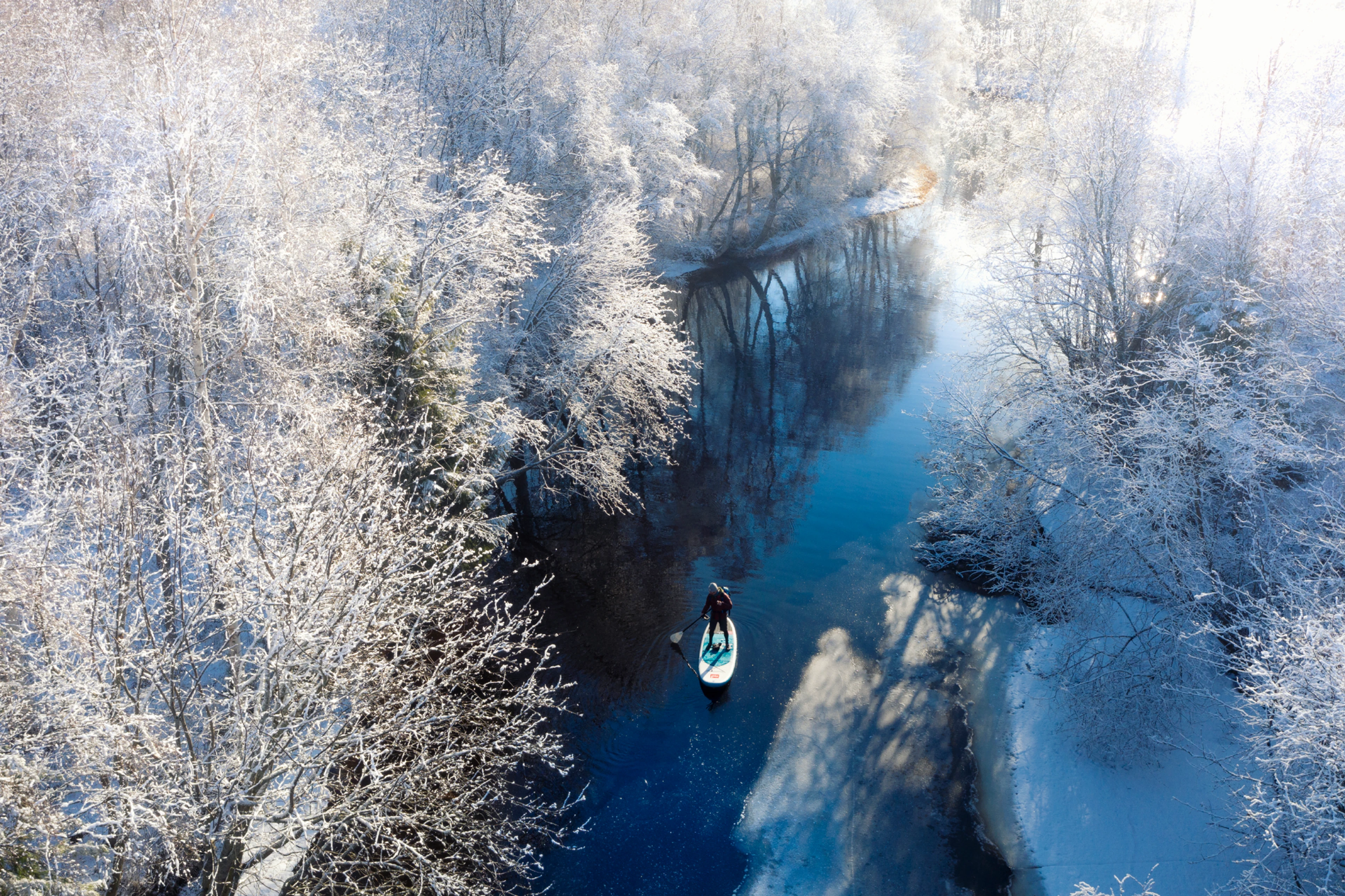Talvinen ilmakuva, jossa henkilö melomassa suppilauta sinisellä joella huurteisten puiden keskellä.