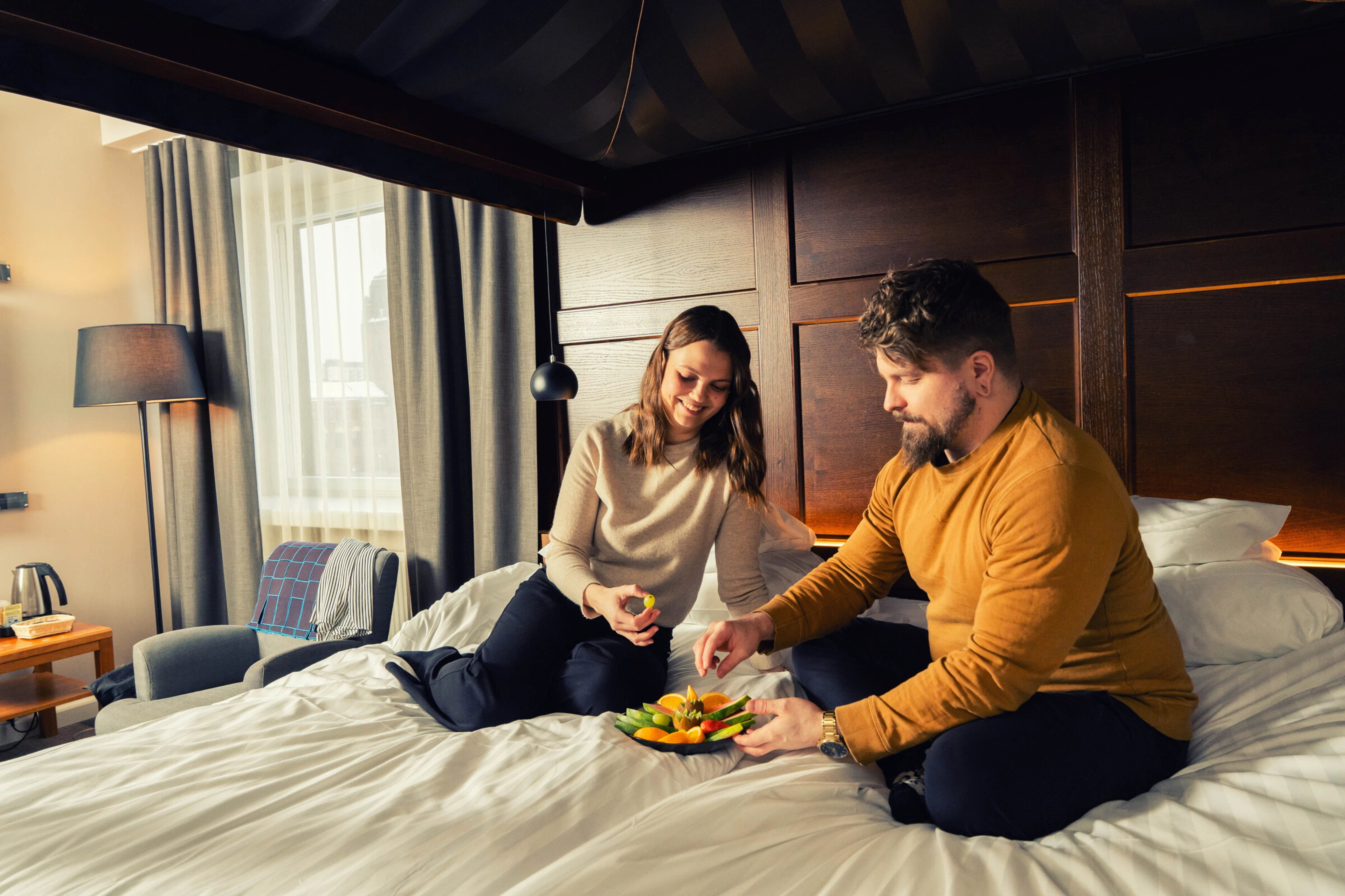 Hotel room with large bed, dark wood paneling and large window with curtains.