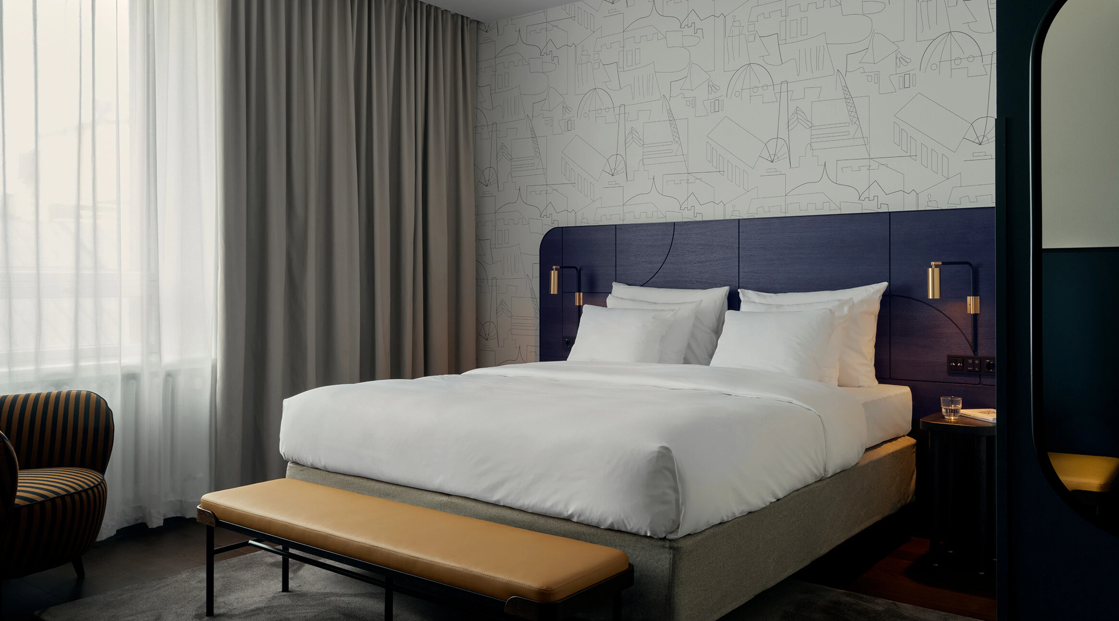 Modern hotel room with a large double bed featuring a dark blue headboard, decorative cityscape wallpaper, and curtains in front of large windows.