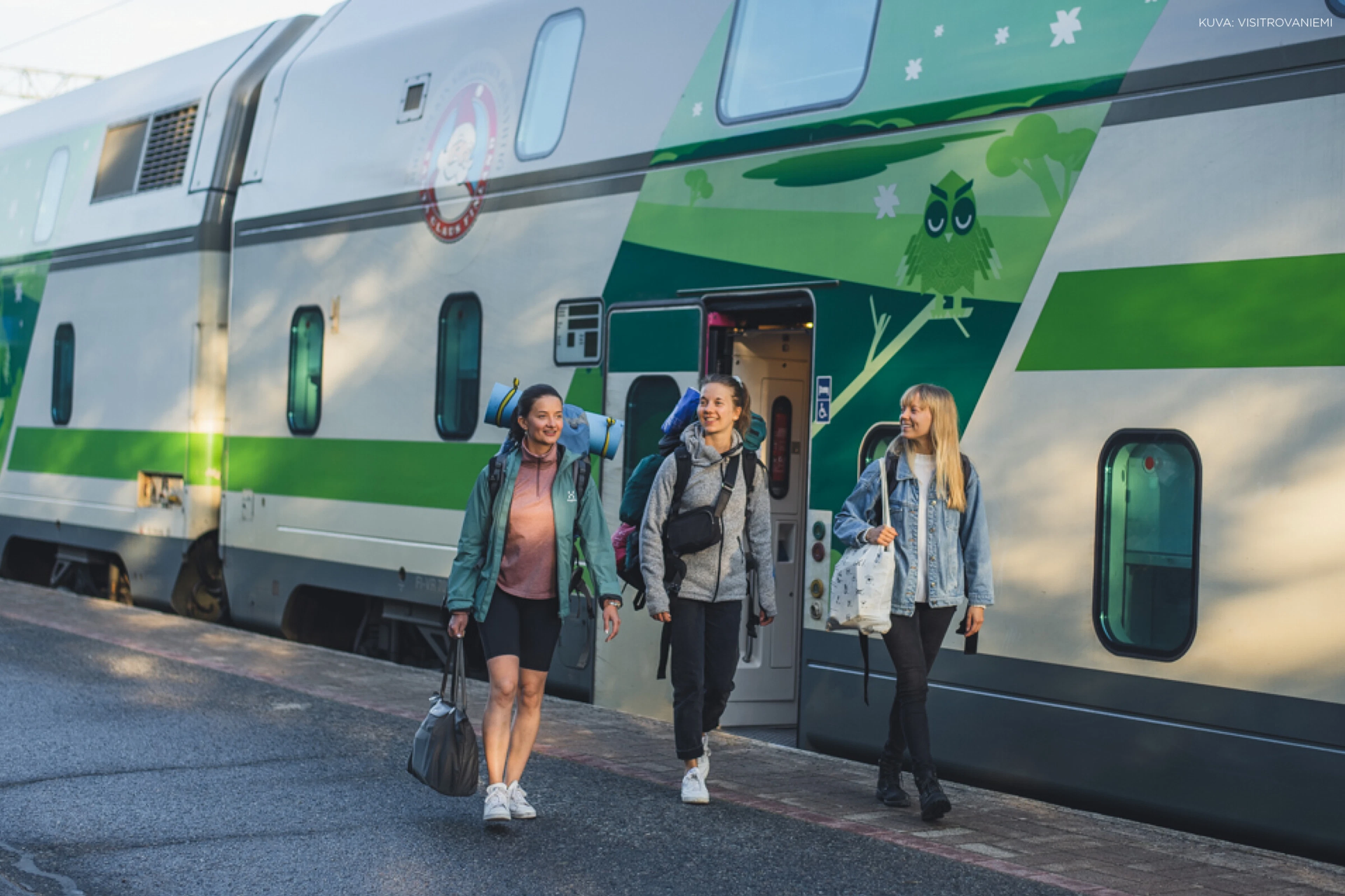Kolme nuorta naista kävelee laiturilla kaksikerroksisen junan vieressä.
