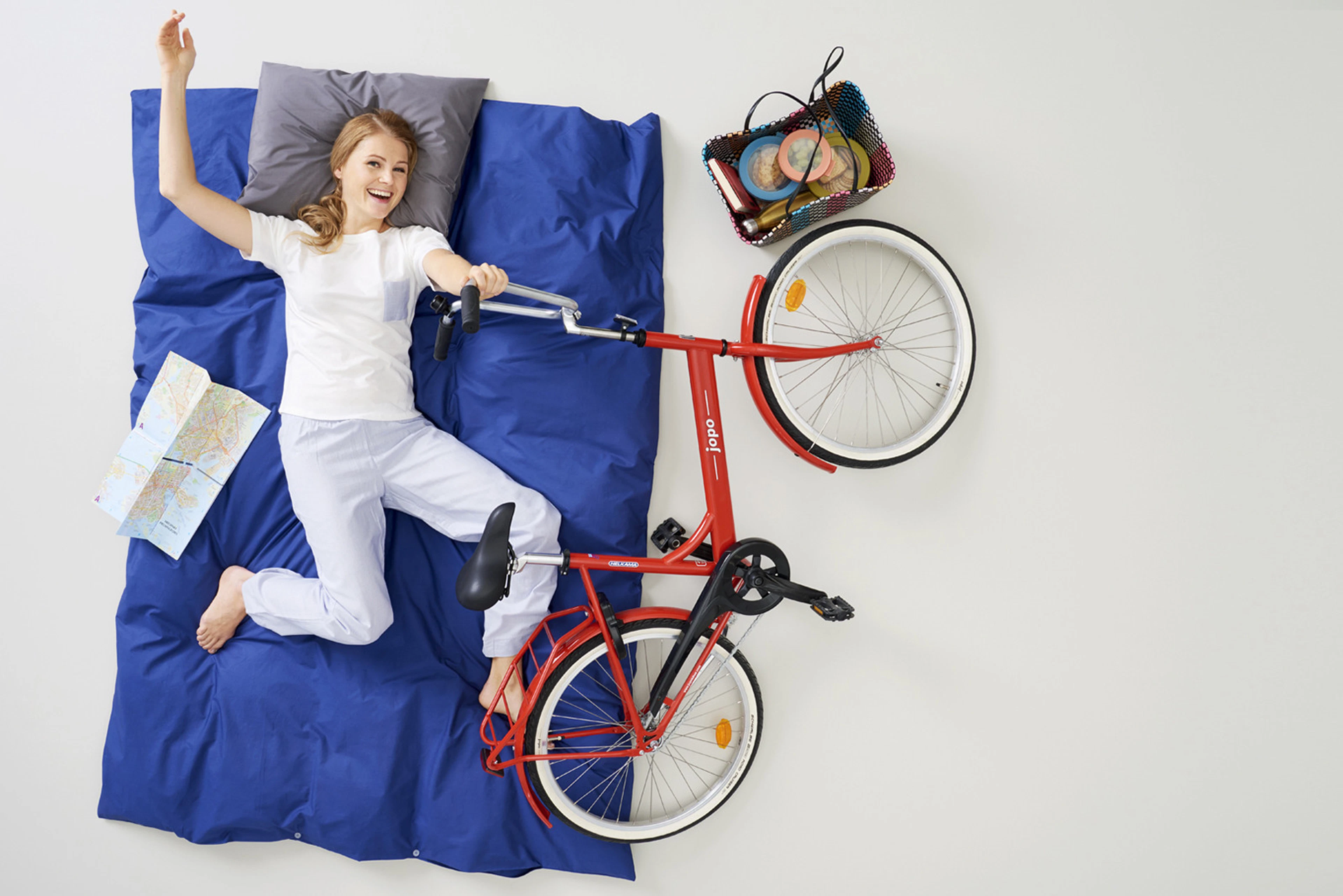 Woman lying on blue bedding with red bicycle beside her, bike basket containing travel supplies.