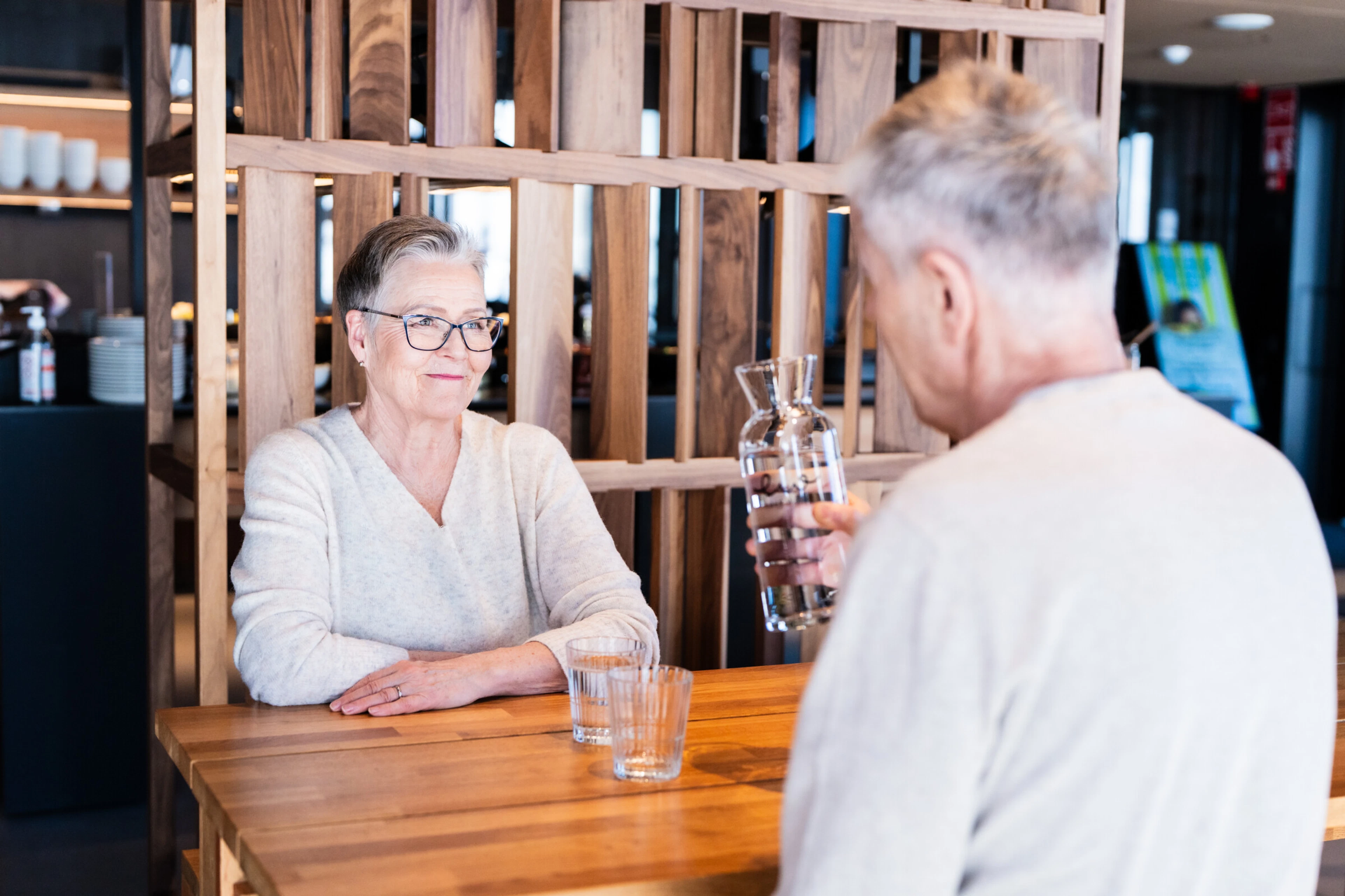 Two people seated at a wooden table beside a slatted wooden screen partition. Even lighting.