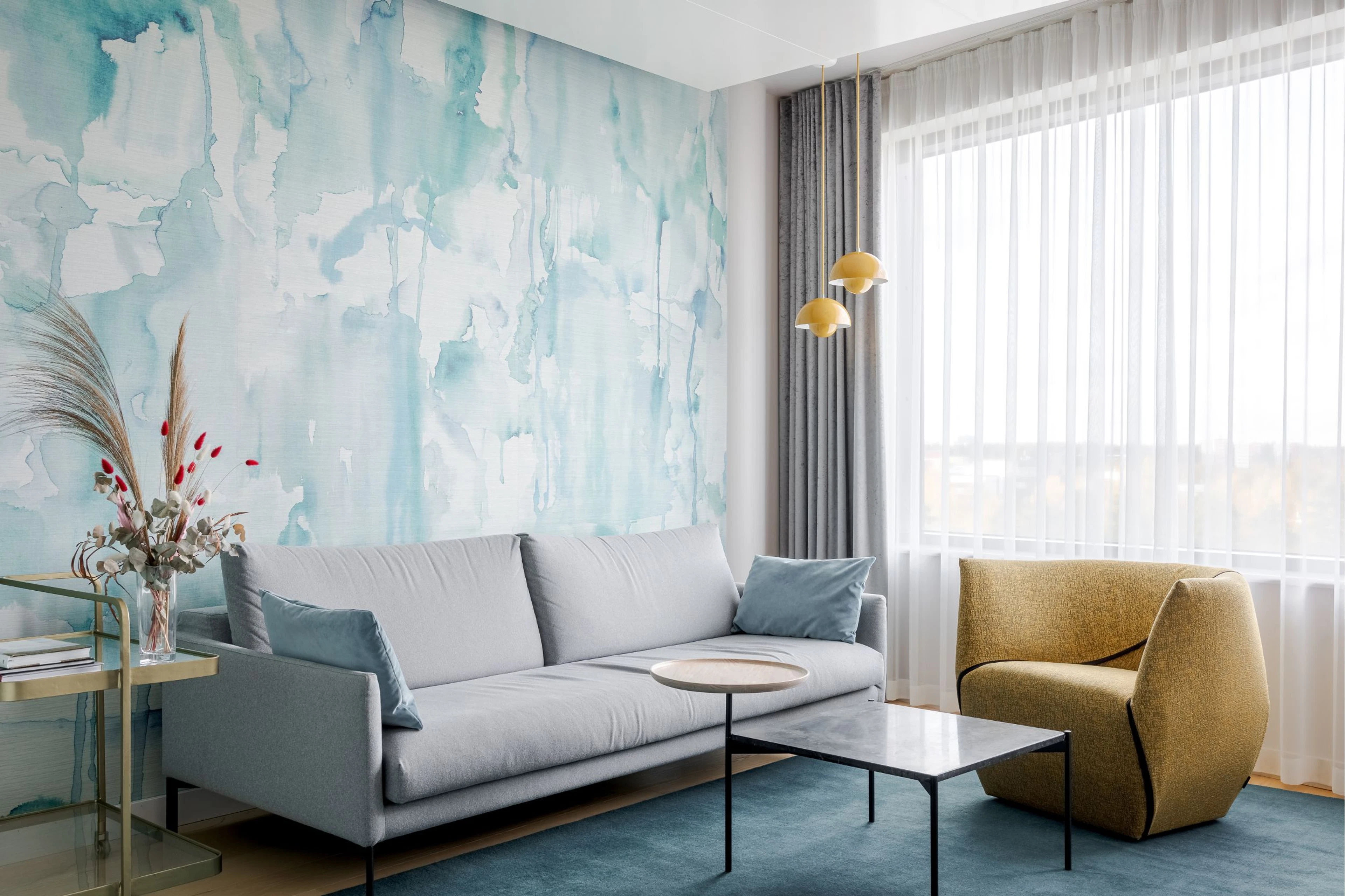 A coffee table surrounded by a wide sofa and a chair next to a window. Behind the sofa there is a bright blue abstract mural wall