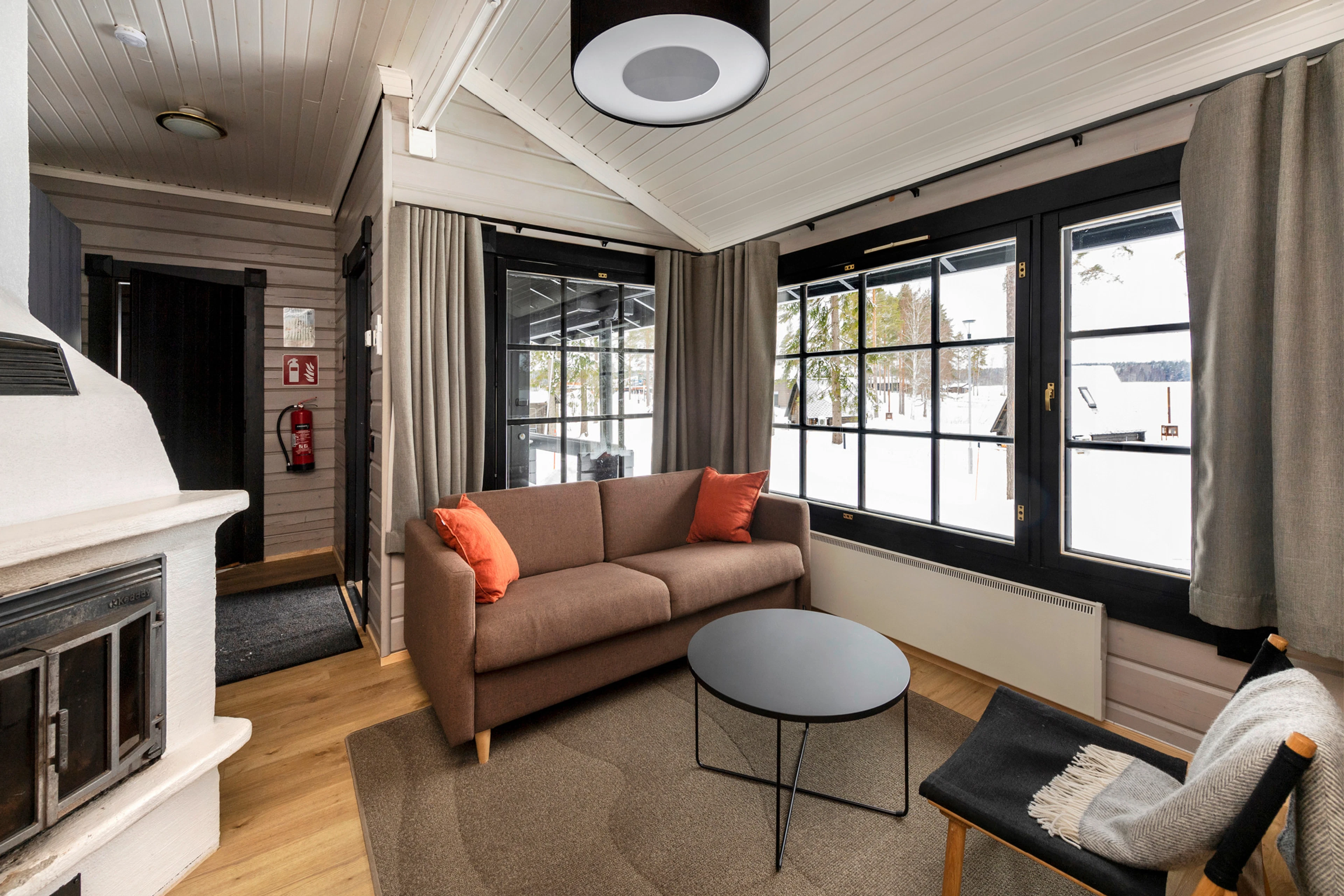 Valoisa hirsimökin olohuone, jossa ruskea sohva oransseilla tyynyillä, puulattia ja suuret ikkunat talviseen maisemaan.