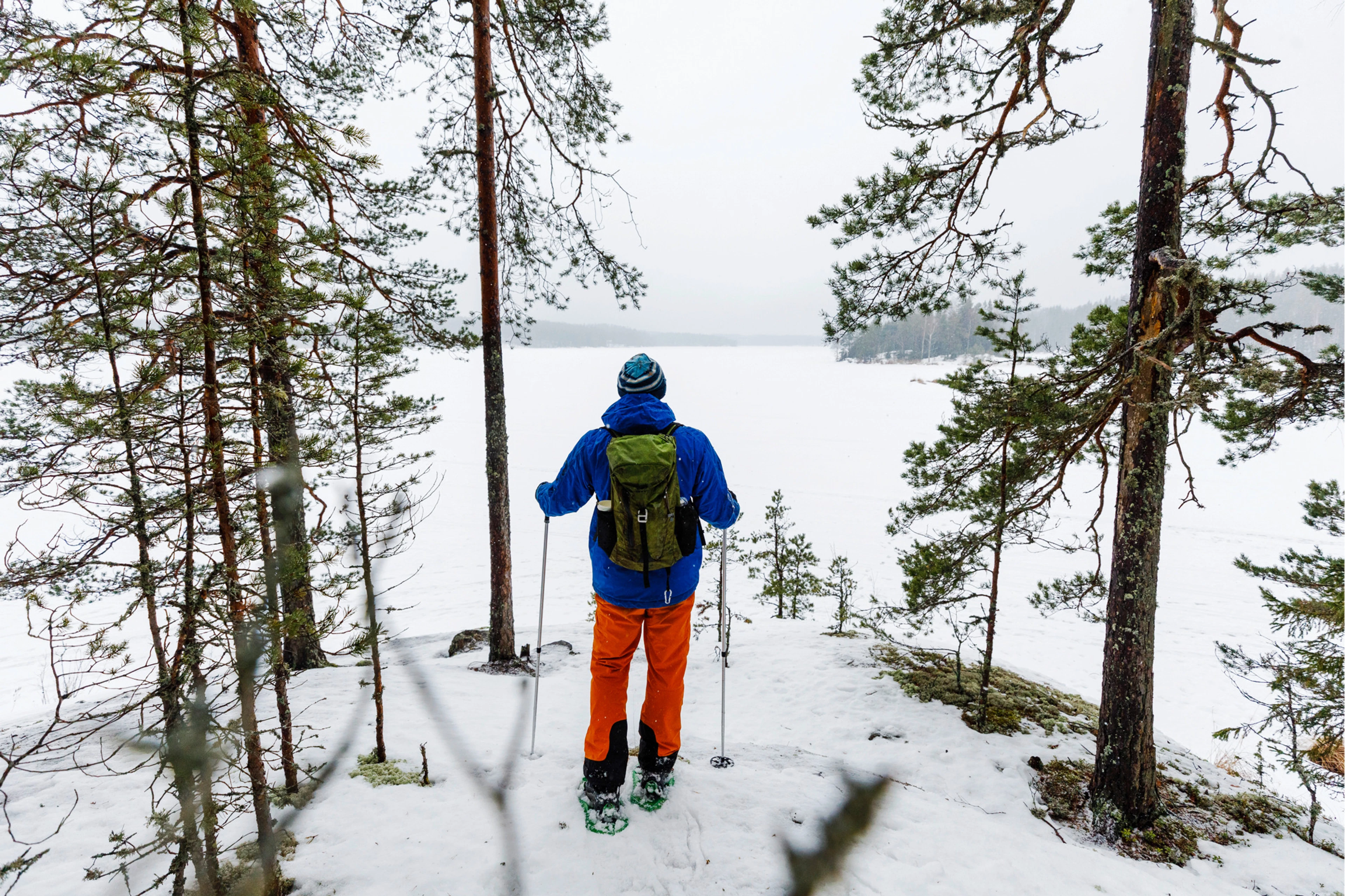 Retkeilijä sinisessä takissa ja oransseissa housuissa lumikengillä mäntymetsän reunalla, katsellen jäätynyttä järveä talvimaisemassa.