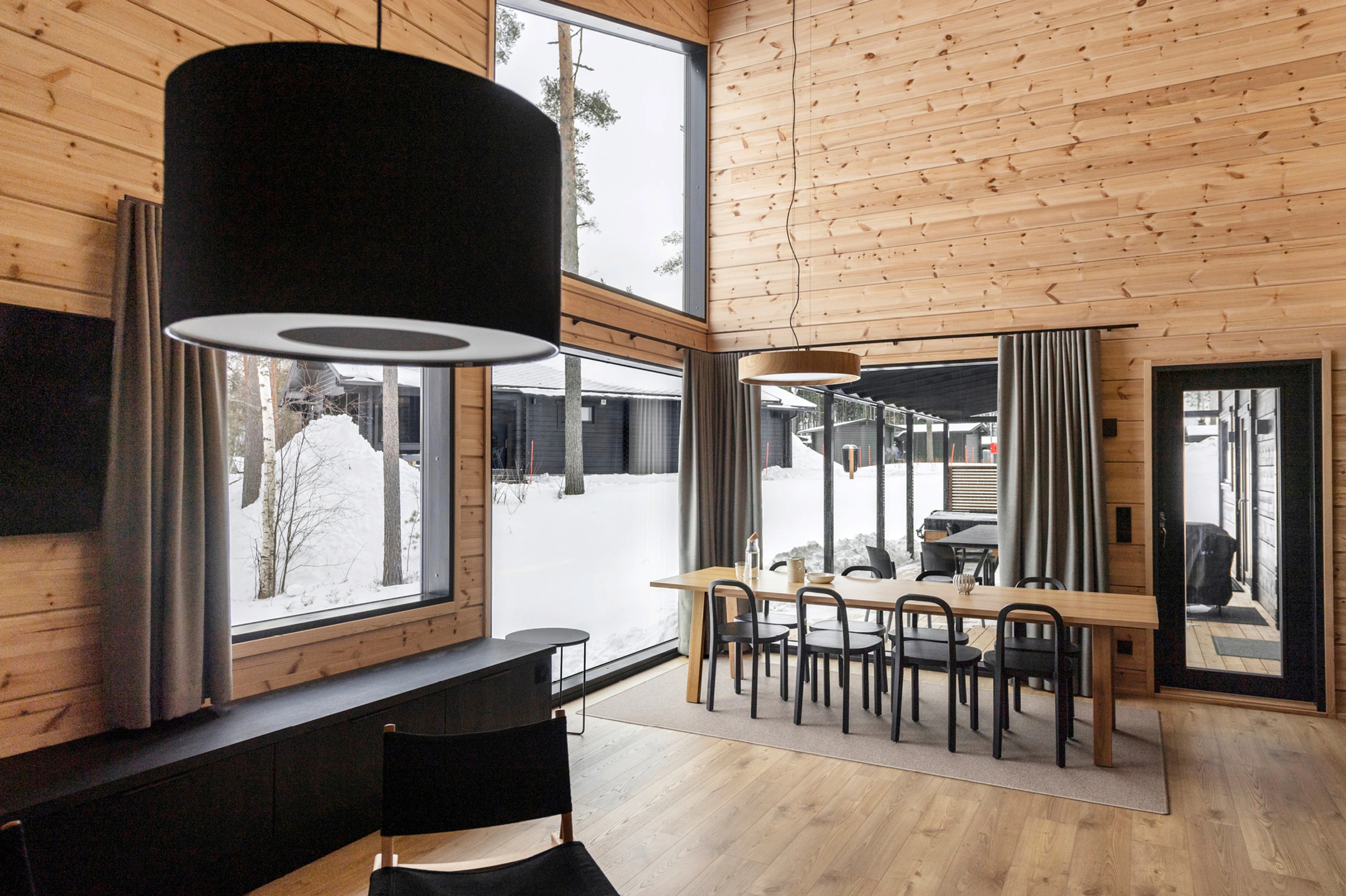 Moderni puupintainen ruokailutila suurilla ikkunoilla, joista näkyy luminen maisema. Vaalea puulattia ja korkea katto.