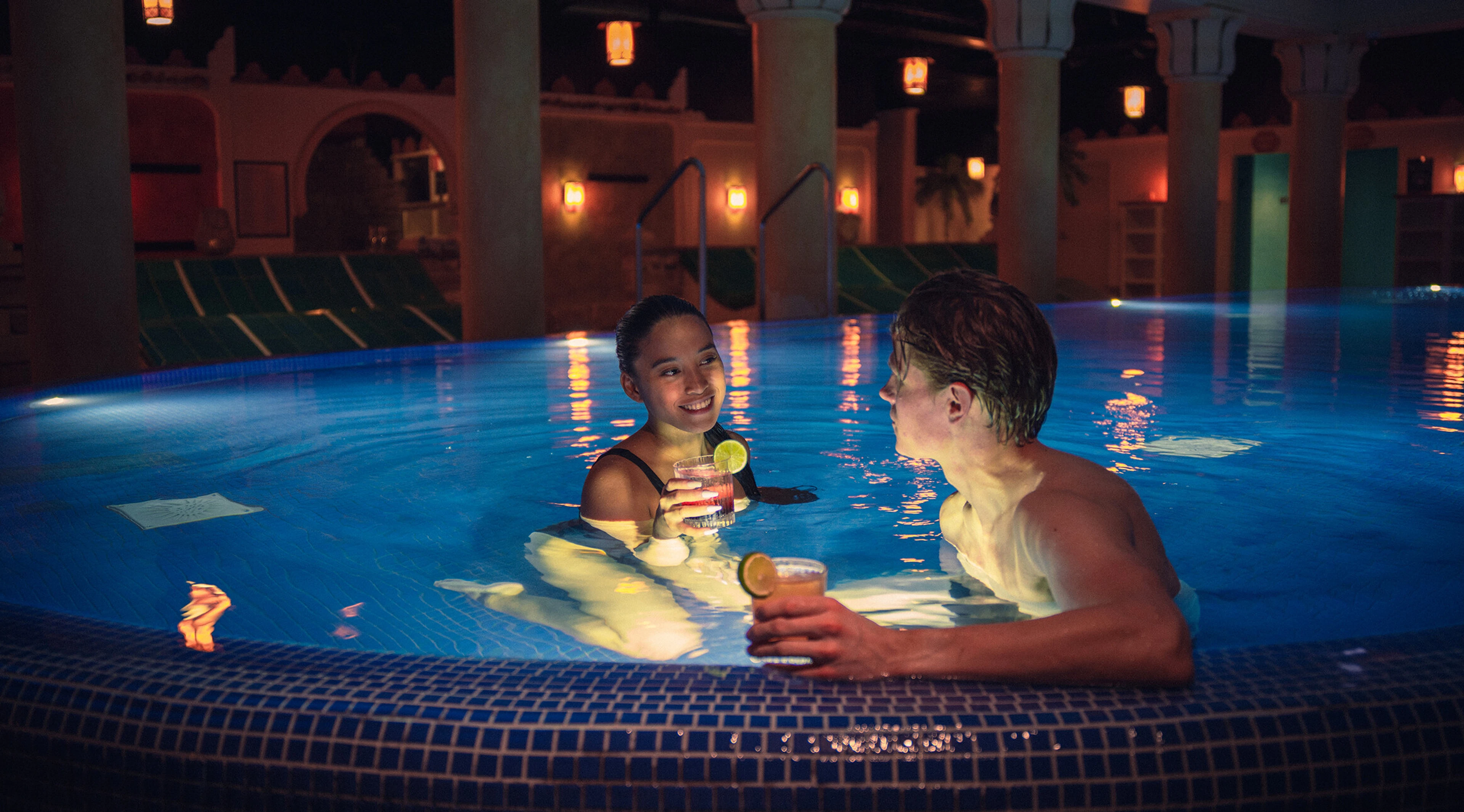 Two people in a pool with a stepped edge. Sun loungers and entrances visible in the background.