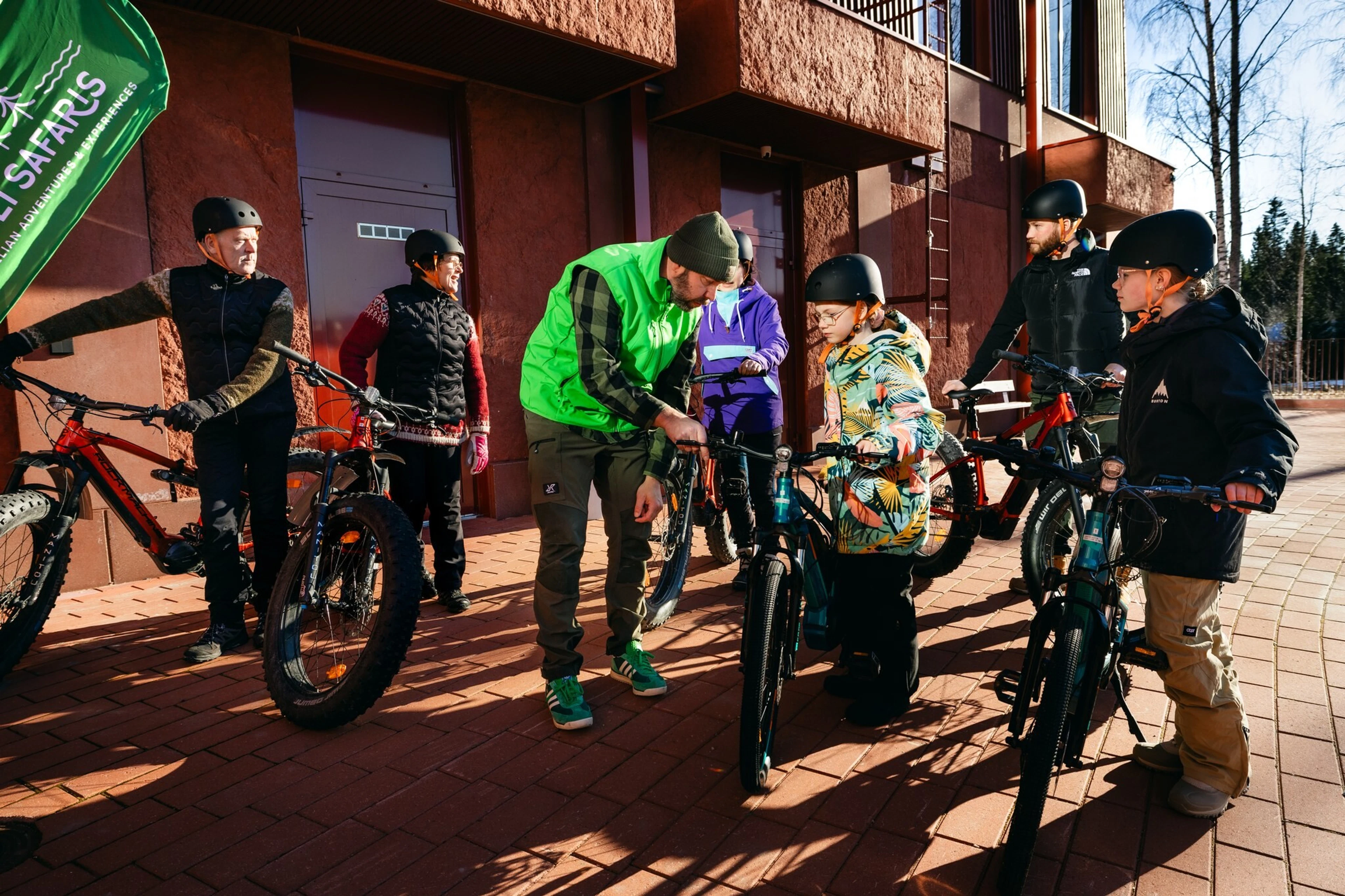 En guide i grön väst visar en cykeldel för människor med hjälm. Gruppen har fatbikes och elcyklar framför en rödbrun byggnad. Underlaget är jämn stenläggning.