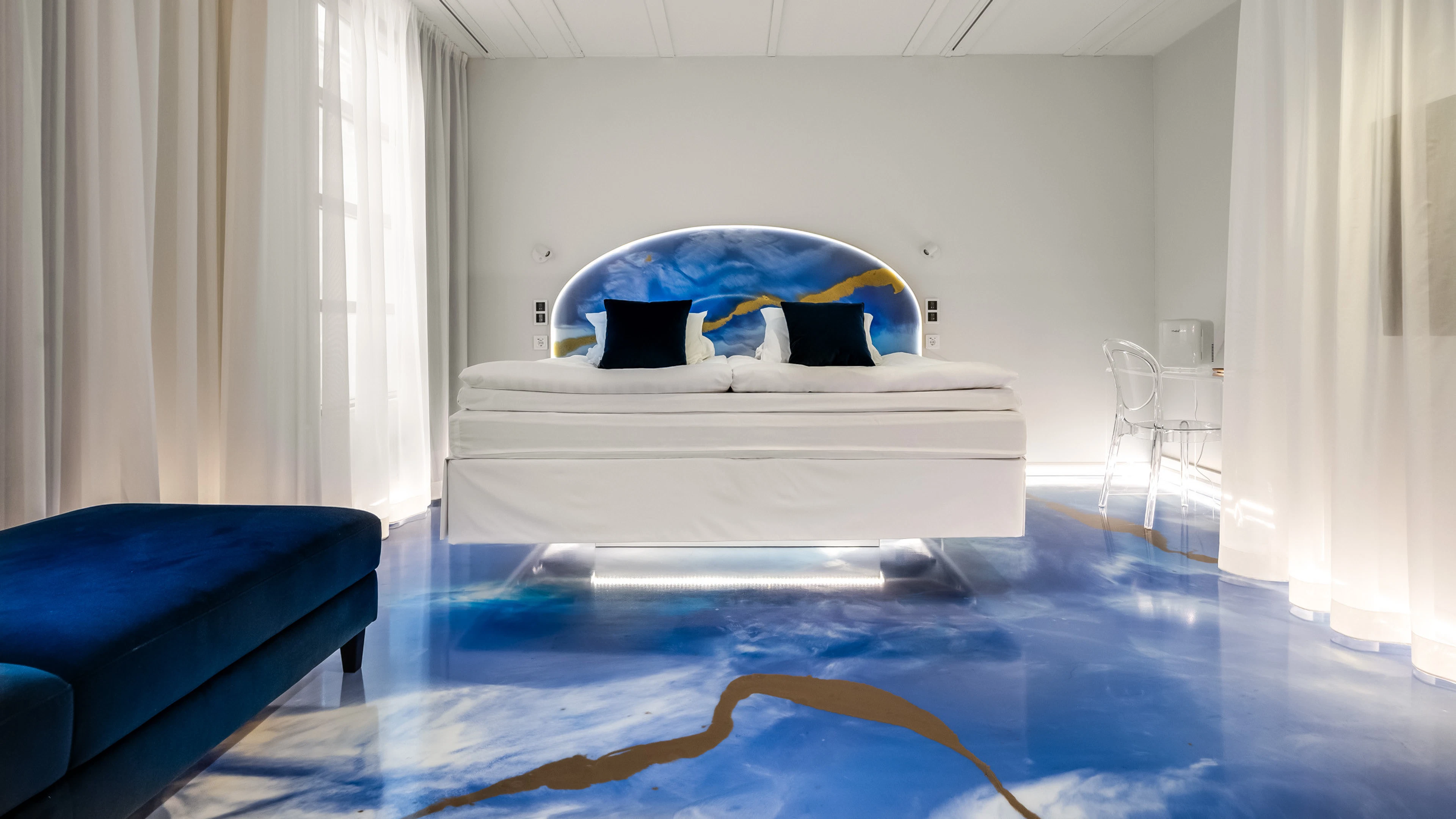 Modern hotel room with white bed and artistic blue floor. Blue sofa on the left, transparent chair on the right.