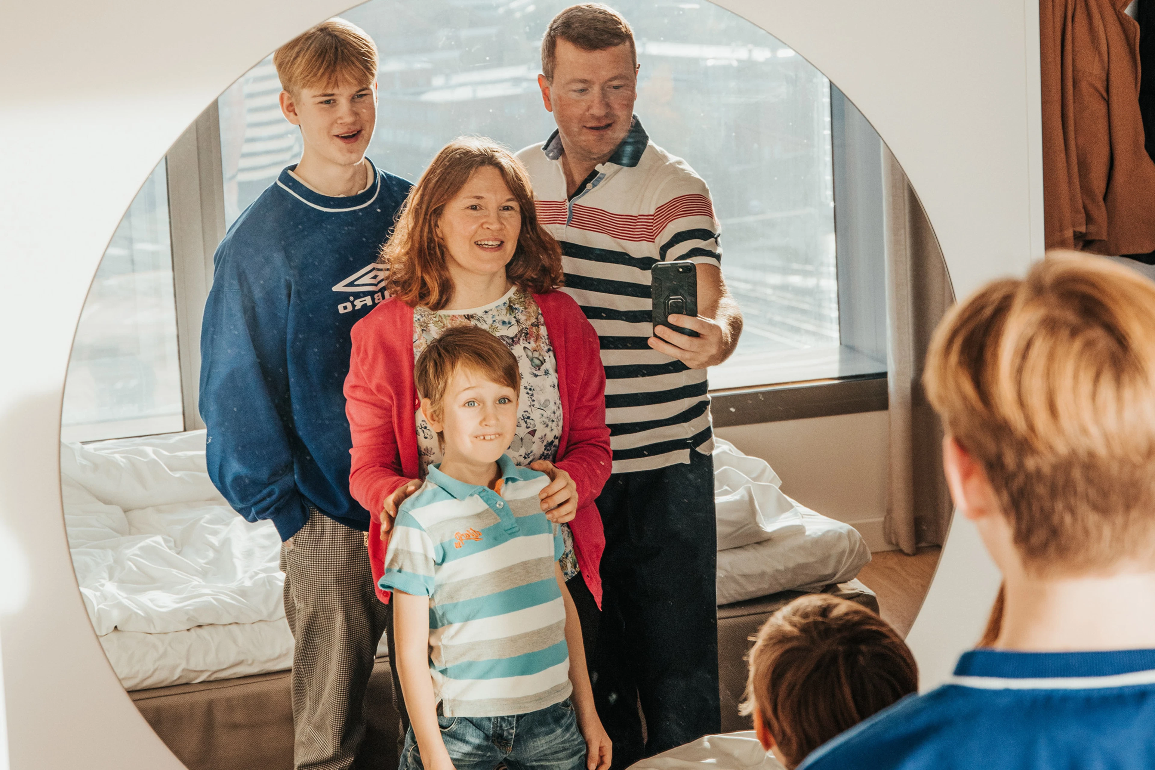 Perhe ottamassa selfietä peilin kautta hotellihuoneessa, jossa on iso ikkuna ja vaalea sisustus.