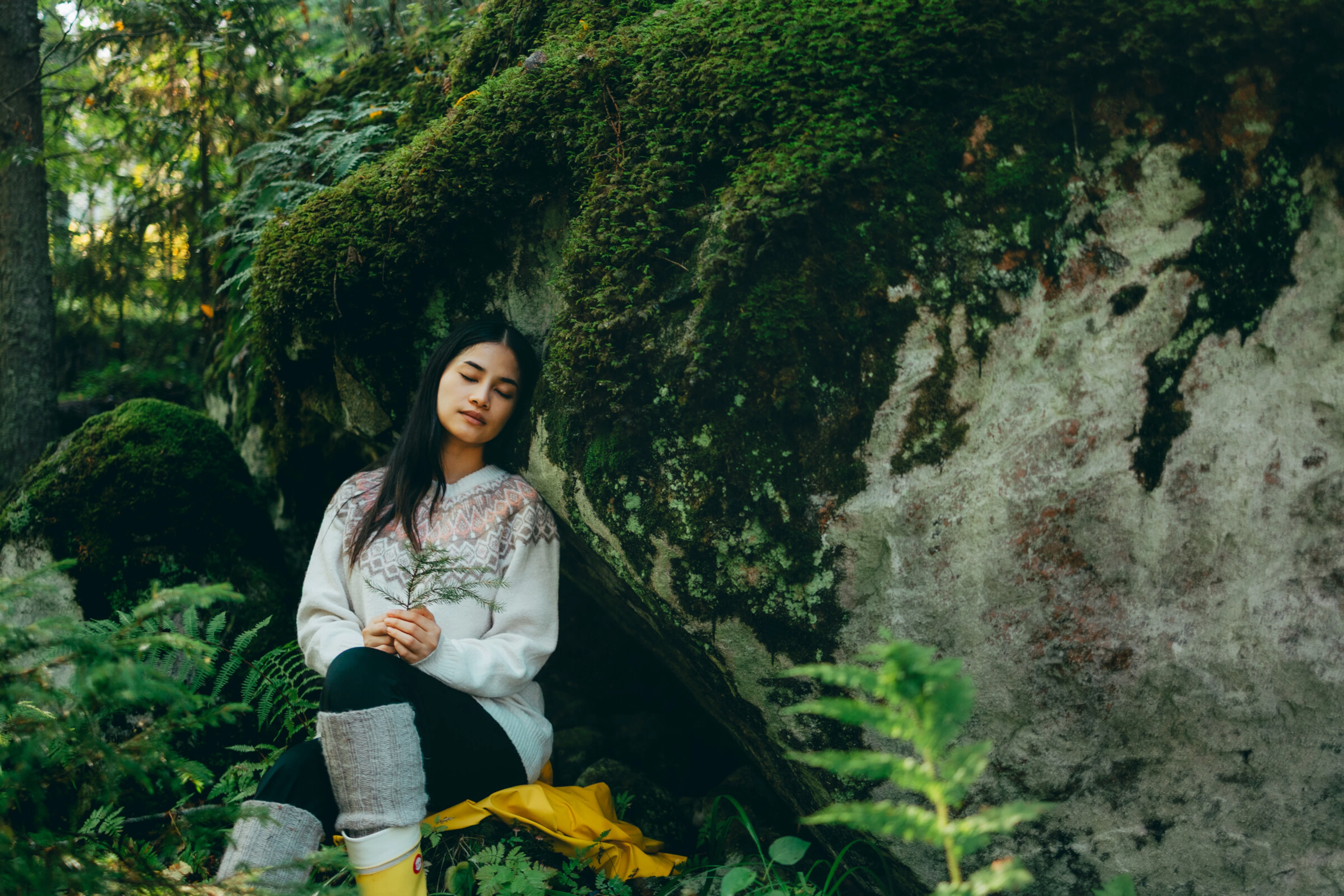 Henkilö istuu sammaleisen kallion vieressä metsässä, yllään valkoinen kirjokuvioitu villapaita ja keltaiset saappaat.