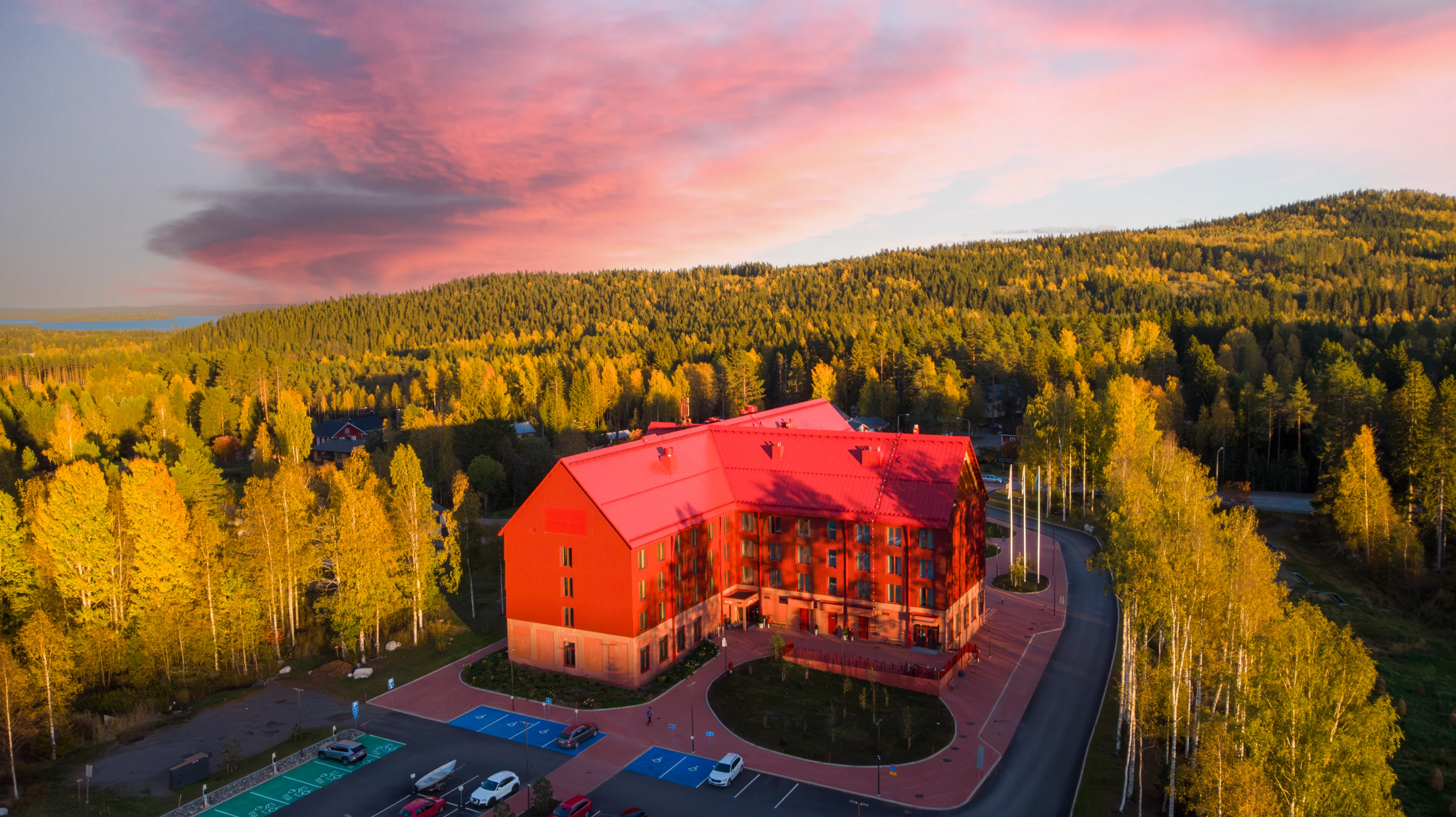 Punainen Kolin Kylähotelli syysruskan värjäämän metsän ympäröimänä, taaempana vaaroja ja taivalla vaaleanpunaisia pilviä.