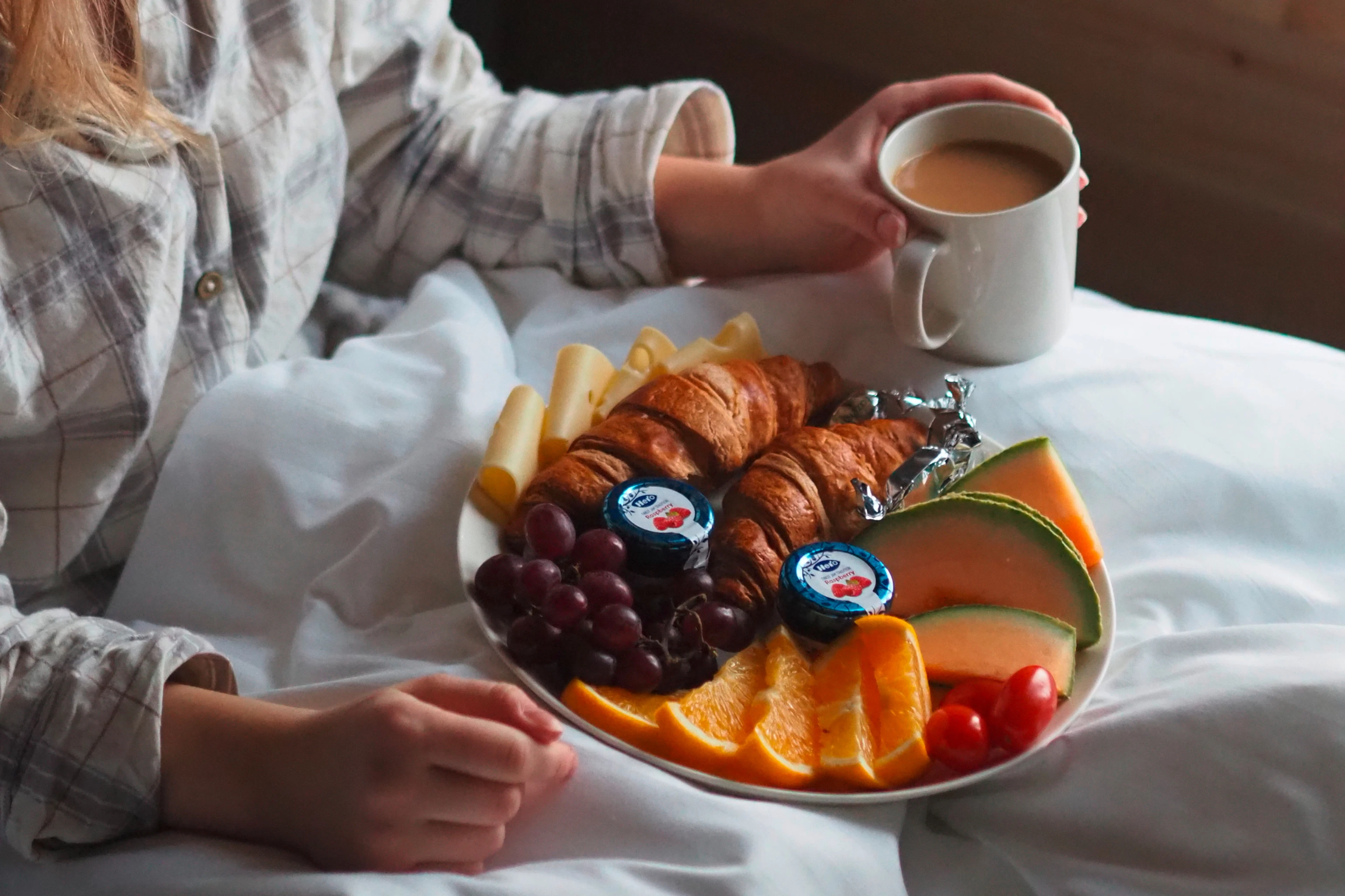 Aamiaislautasella croissanteja, juustoa, viinirypäleitä, melonia, appelsiinia ja tomaatteja. Henkilö pitelee kahvikuppia.