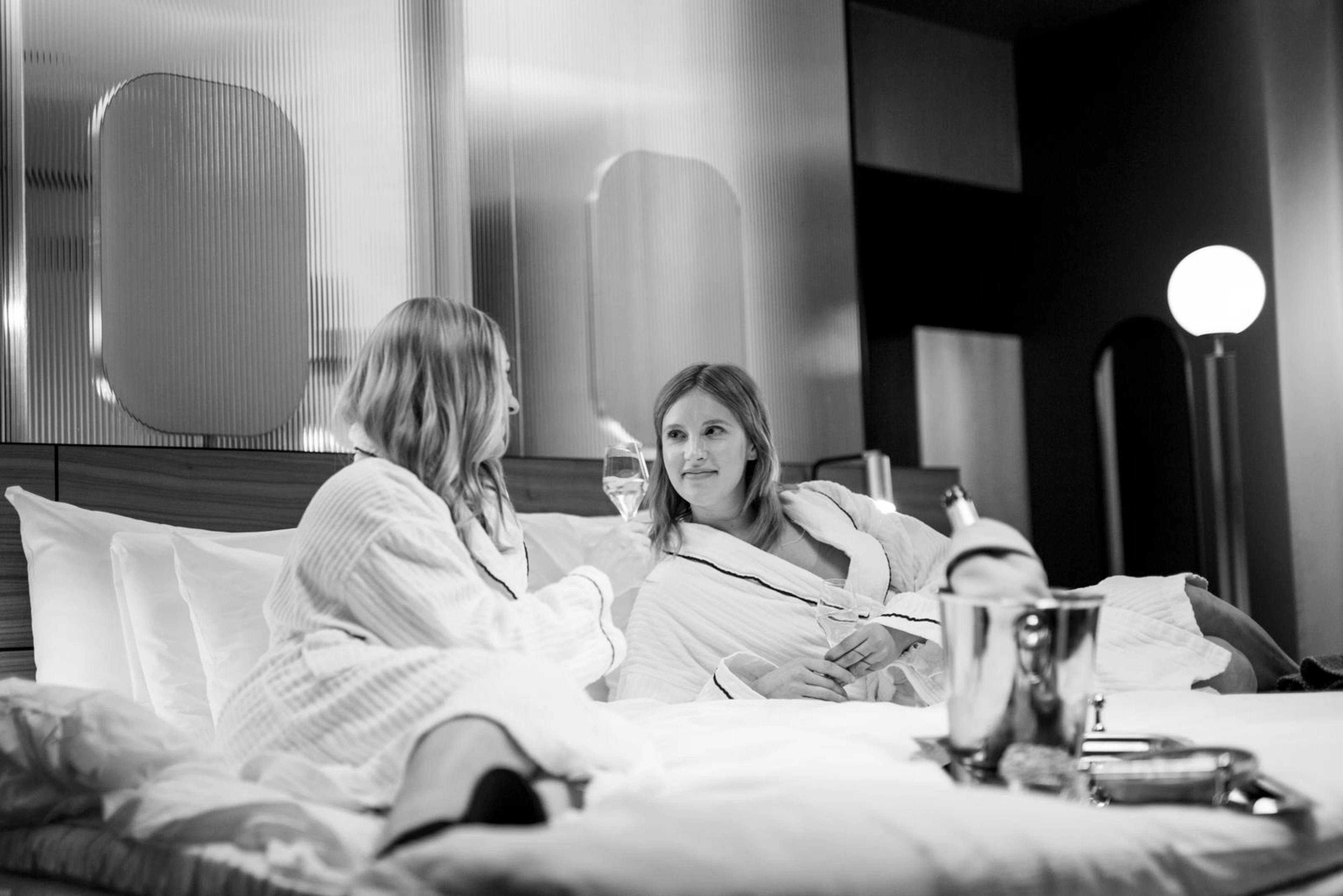 Two people conversing while sitting on the bed in a modern hotel room. The bed is surrounded by level flooring.