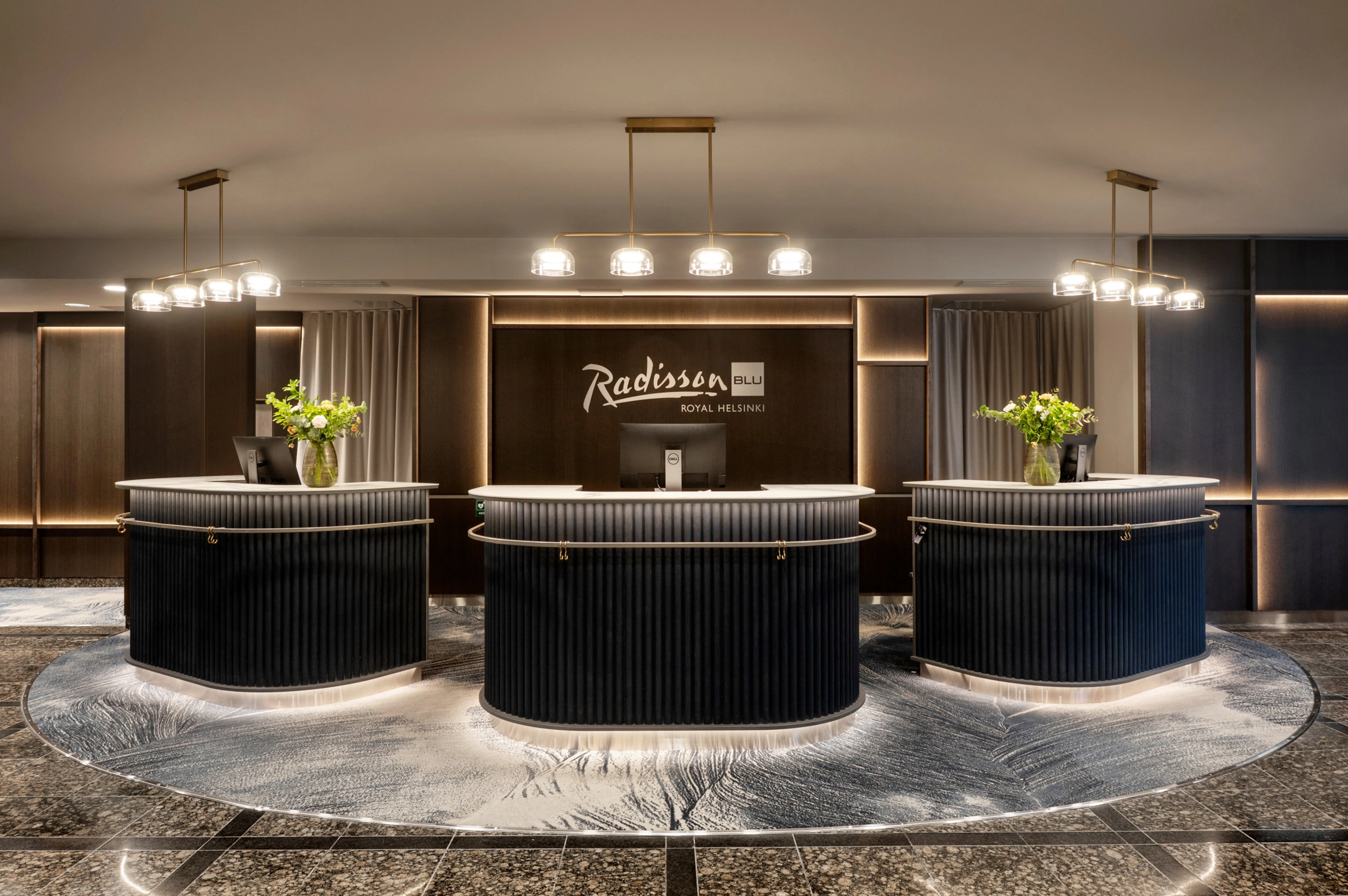 Elegant hotel reception area with three curved desks, floral arrangements and stylish pendant lighting. Smooth marble flooring.