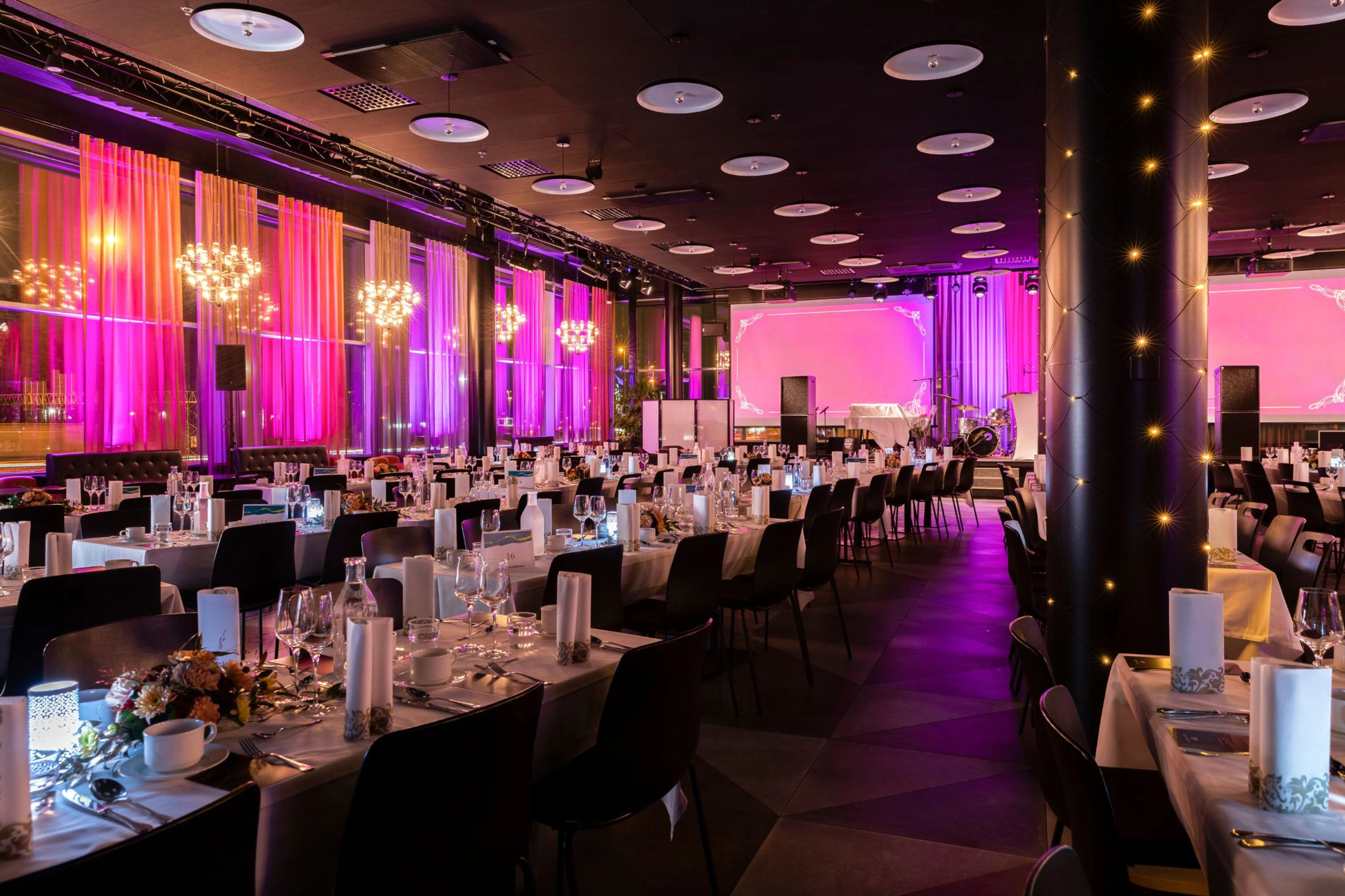 Banquet hall with white tablecloths under pink lighting. Space features sheer curtains, decorative lights and a performance stage.