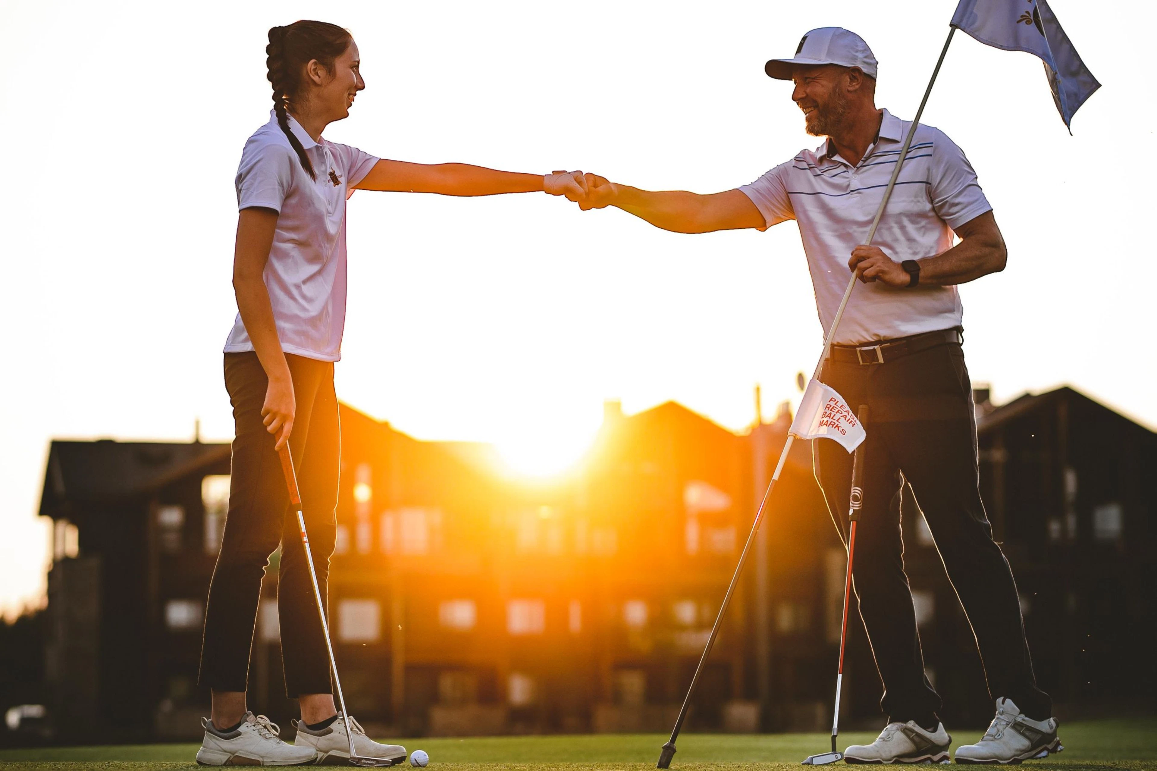 Golfaajat lyövät kättä golfkentällä auringonlaskussa. Takana alppitalojen takaa kajastaa aurinko.