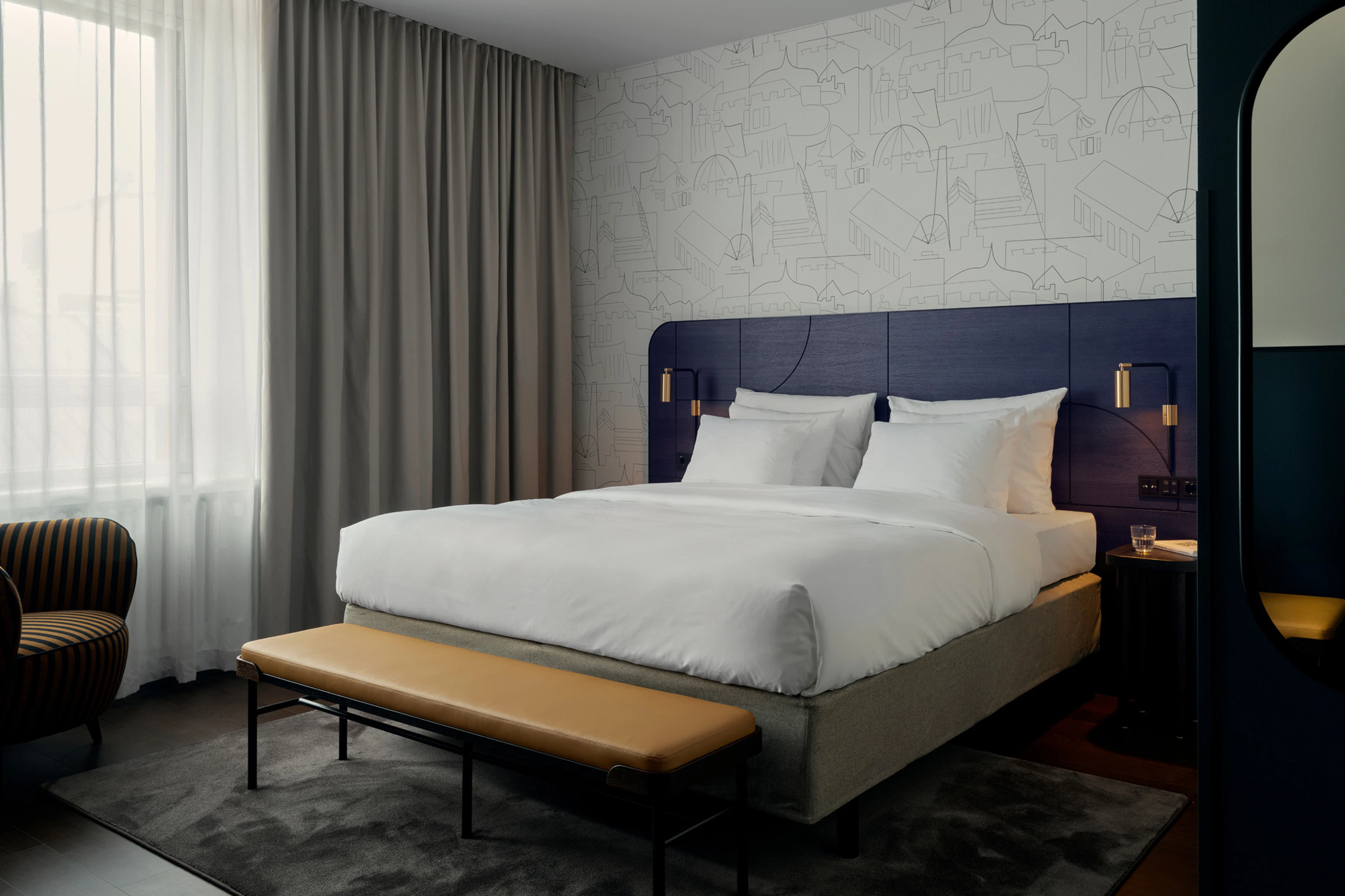 Tyylikäs hotellihuone, jossa parivuode tummansinisellä päätypaneelilla, vaaleat lakanat ja nahkainen penkki. Seinällä linjapiirrostapetointi.