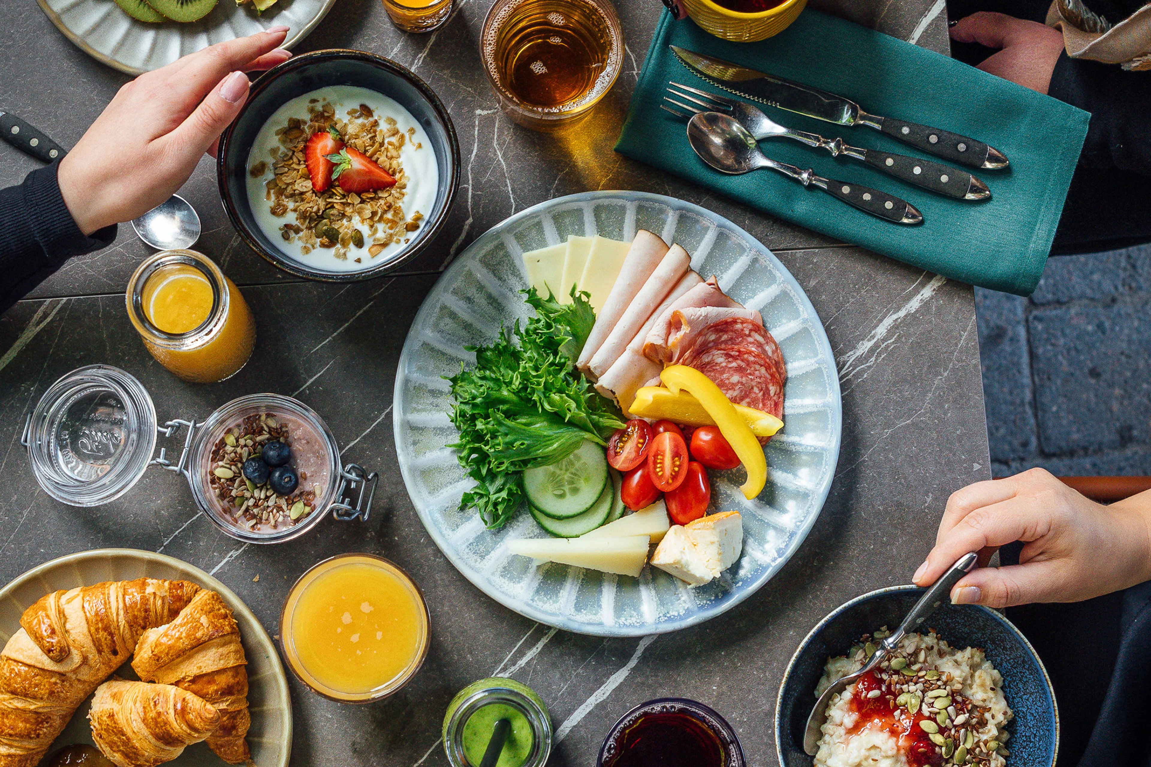 Abundant breakfast spread on marble table featuring a plate with meats, cheese, vegetables, yogurt bowls with granola, croissants, and glasses of juice.