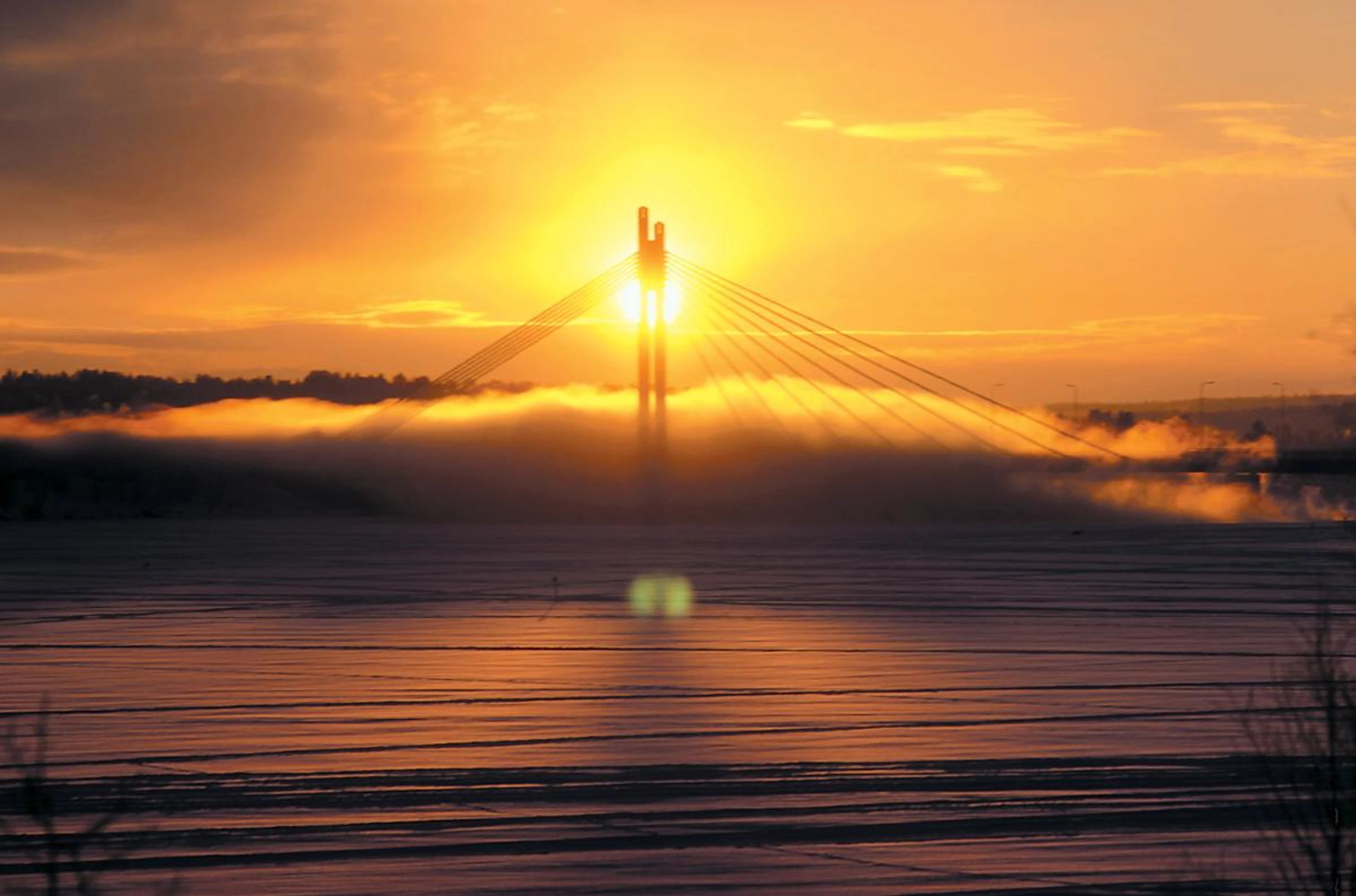 Silta auringonlaskussa, aurinko paistaa suoraan sillan pylväiden takaa, ympärillä oranssia taivasta ja usvaista maisemaa.