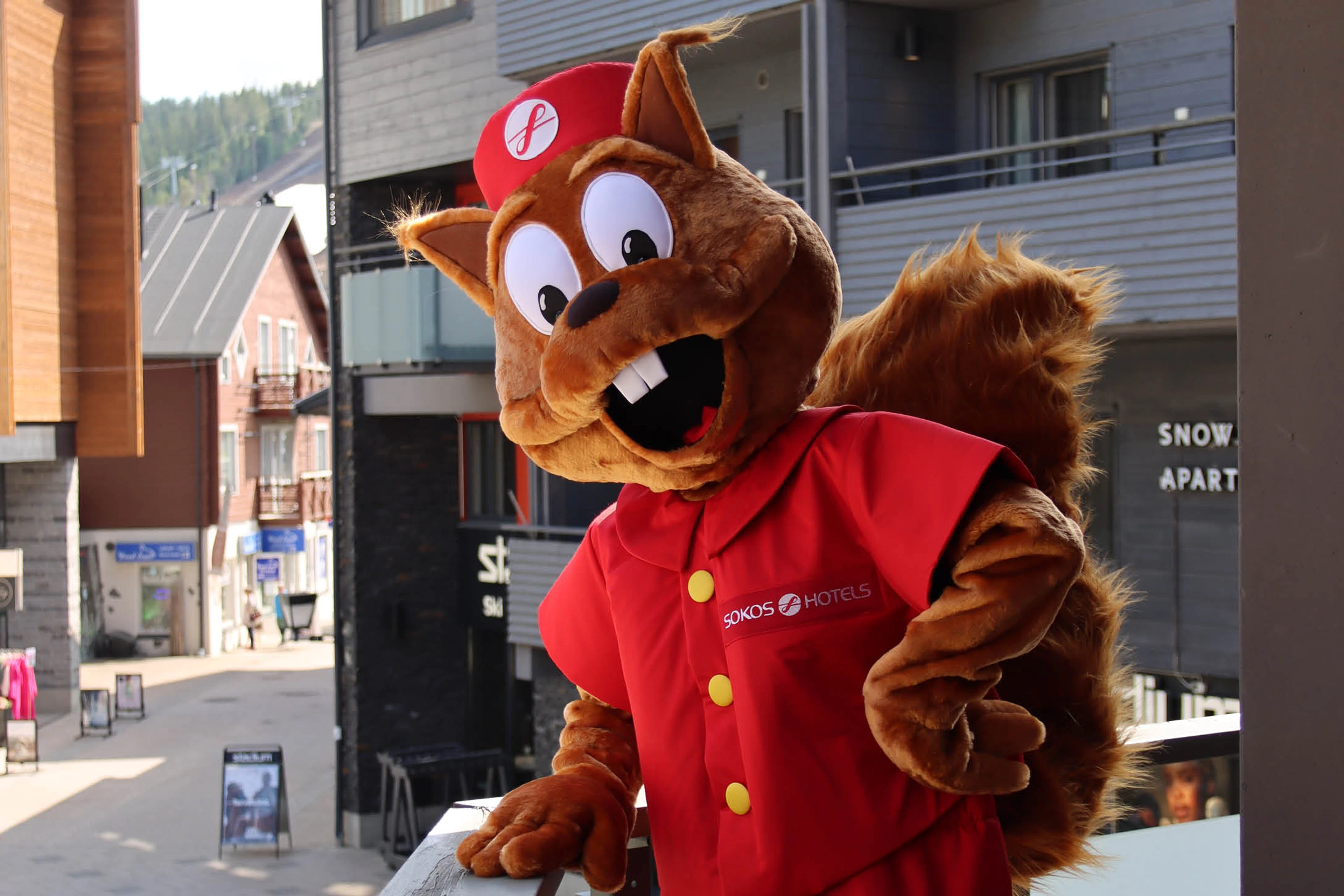 Brown squirrel mascot in red uniform and cap posing outdoors in front of a hotel.