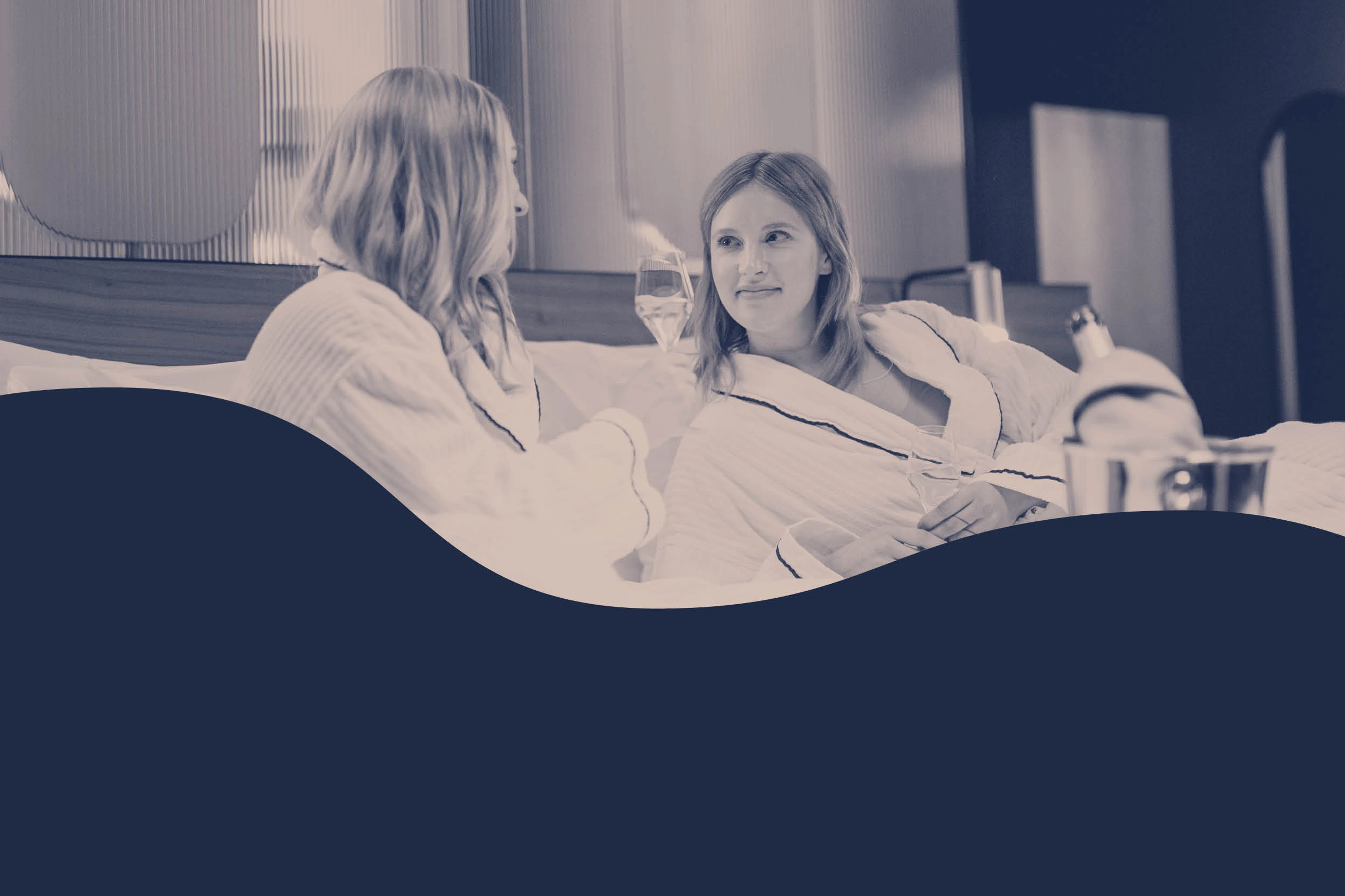 Kaksi henkilöä kylpytakeissa rentoutumassa ja nauttimassa juomasta hotellihuoneessa.