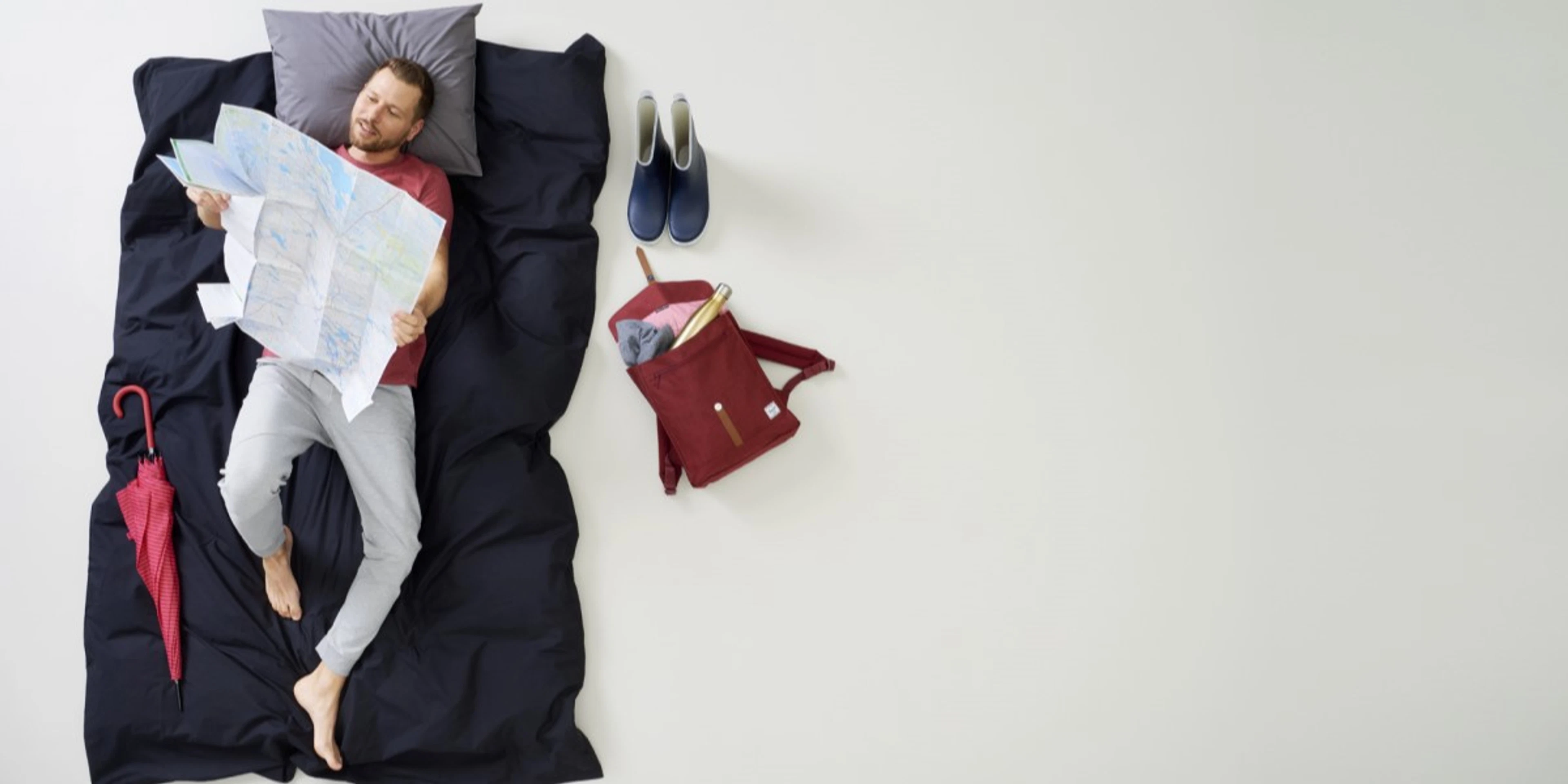 Person lying on a dark blue mattress on a light floor, beside blue shoes, a red umbrella and a burgundy backpack.