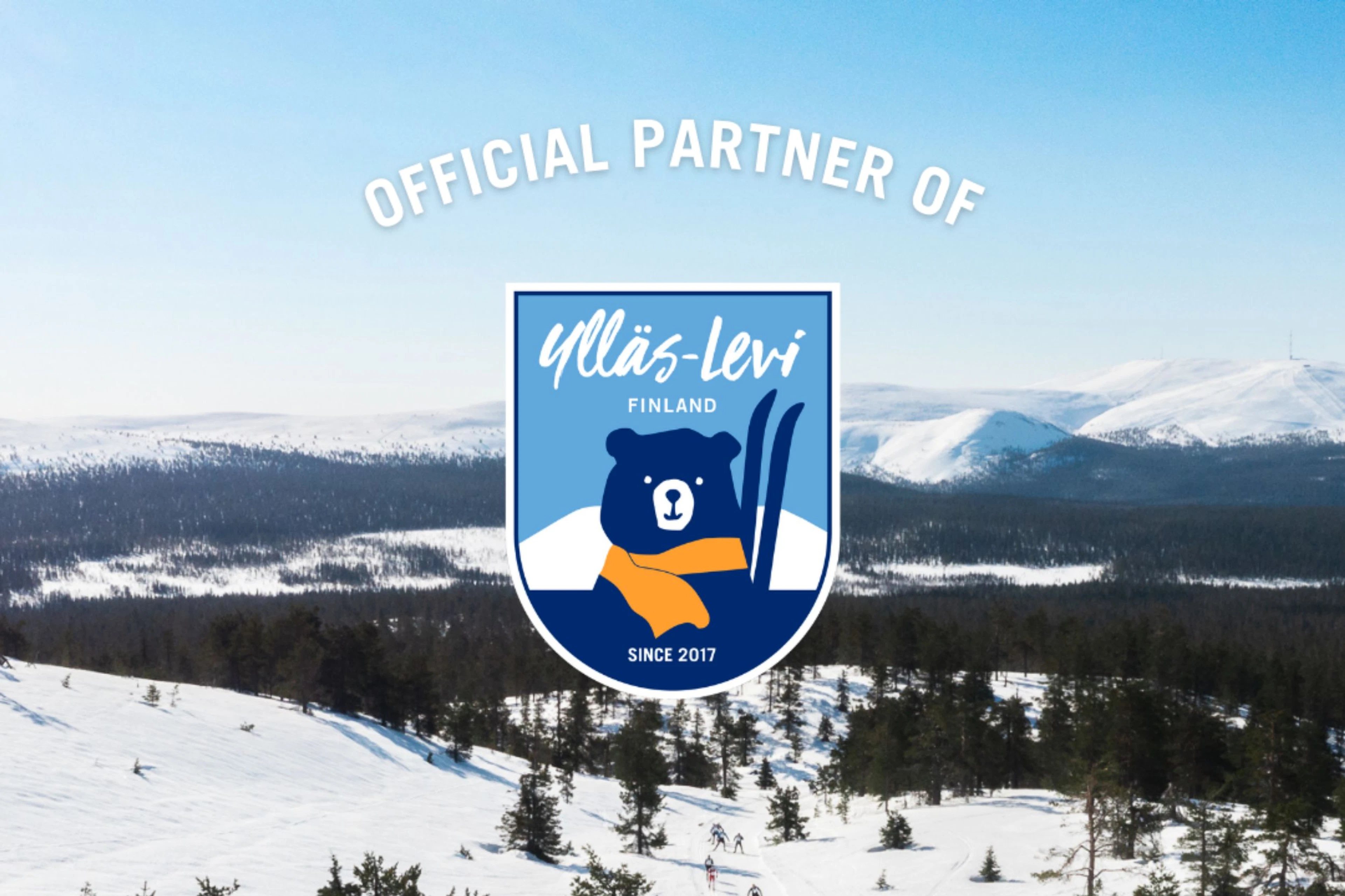 Ylläs-Levi suusakeskuse talvine maastik lumiste mägedega, metsaga ja suusaradadega. Keskel on Ylläs-Levi logo.