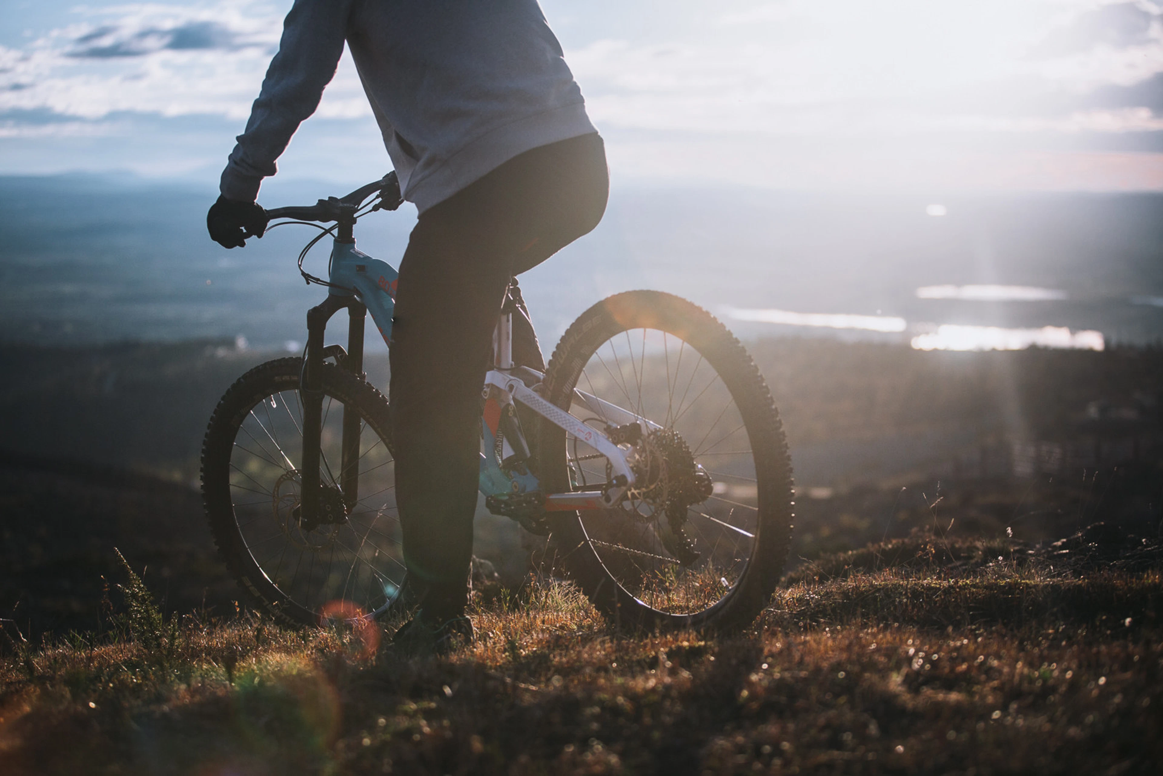 Cyclist on a mountain bike on a grass-covered trail, sunlight in background and landscape at horizon.