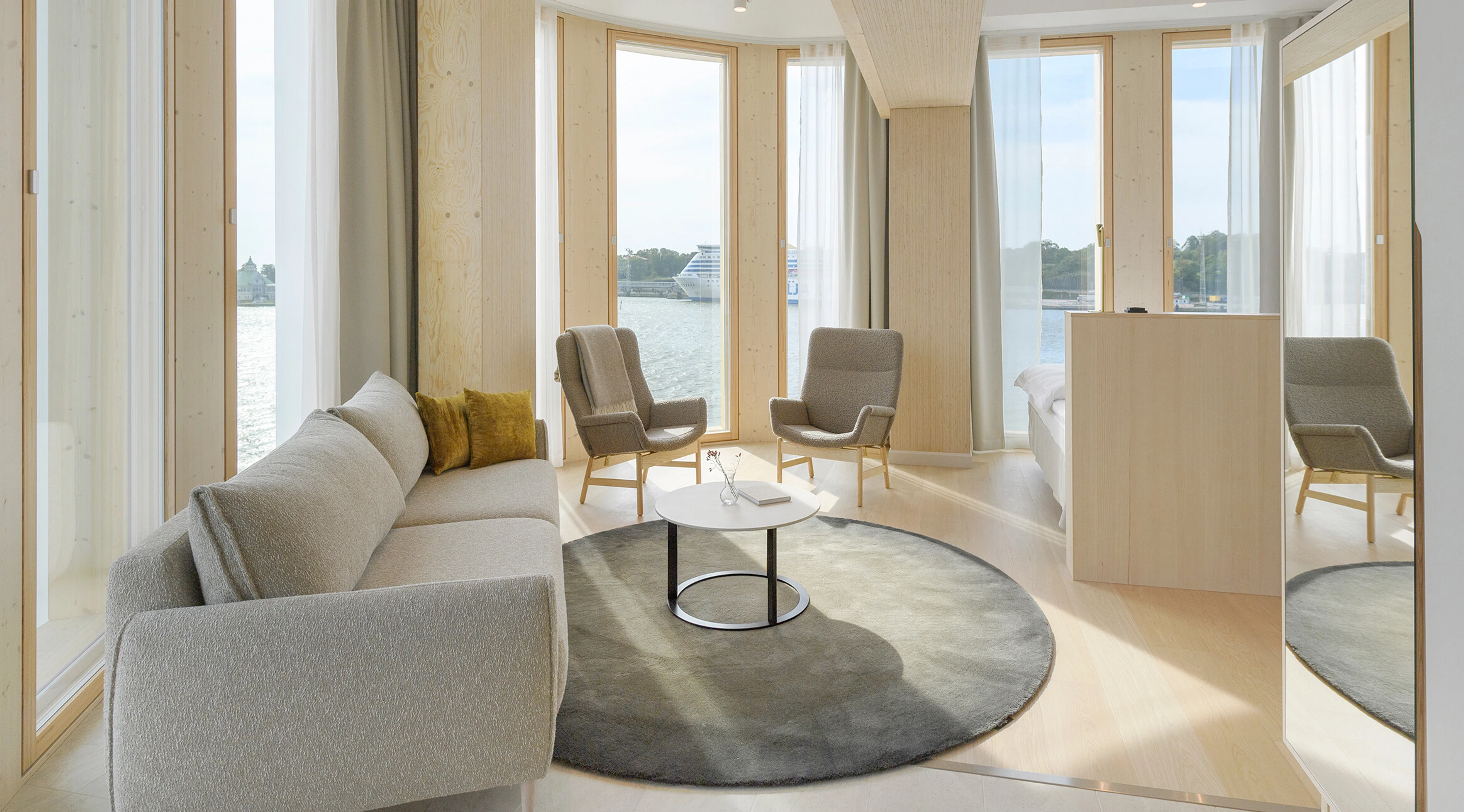 Ljust loungeutrymme med stor rund matta på hårt golv och panoramafönster mot havet.