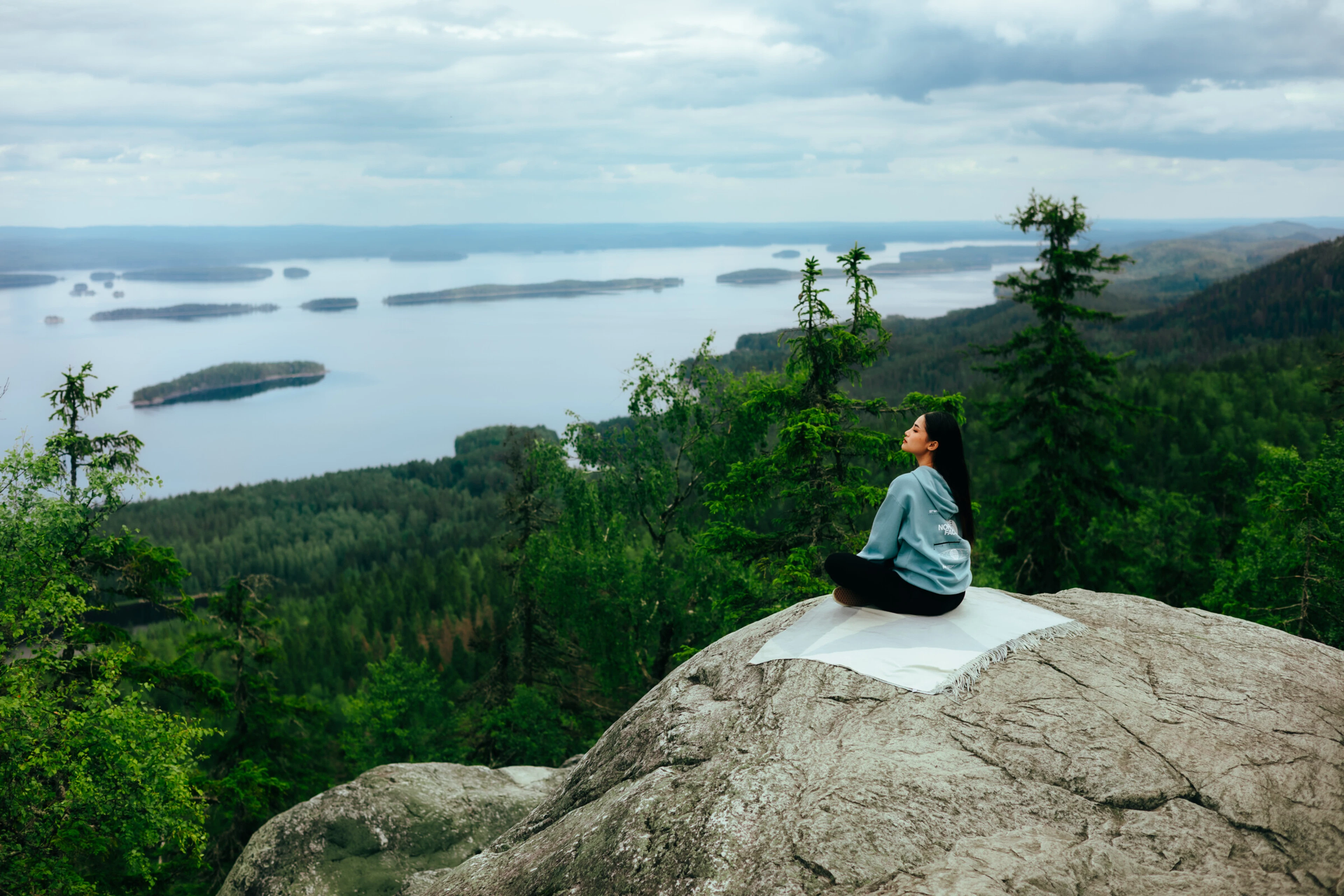 Henkilö istuu Ukko-Kolin kallion huipulla ja katsoo Pielisen järvimaisemaa. Taustalla näkyvät Kolin metsäiset vaaramaisemat ja järvenselkää saarineen sekä pilvinen taivas.